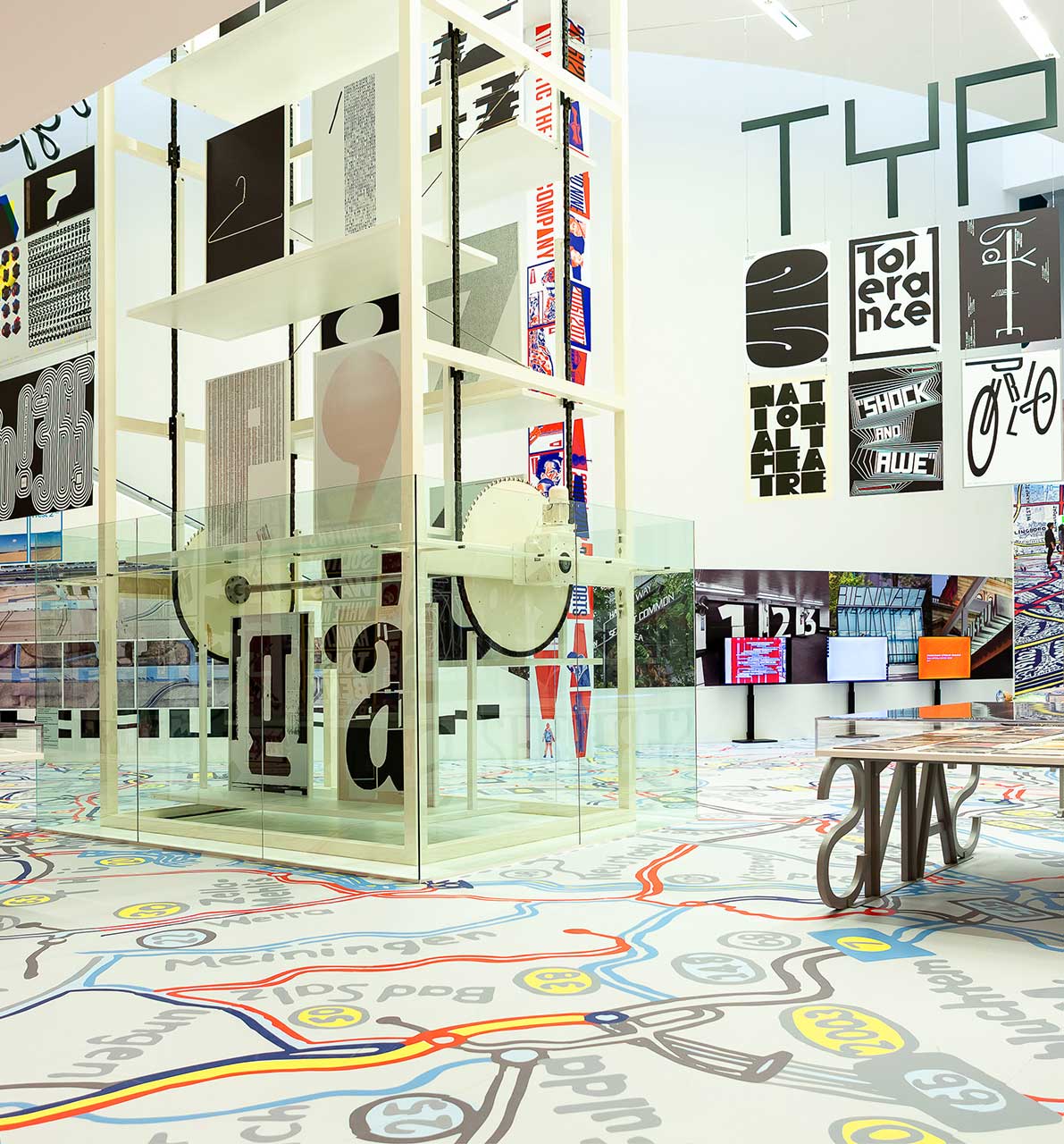 Fotografie Ausstellungsansicht. Der Paternoster (Aufzug) mit Plakaten aus der Ausstellung. Rechts davon eine Vitrine. Im Hintergrund Bildschirme und weitere Poster. Auf dem Boden befindet sich eine bunte Landkarte.