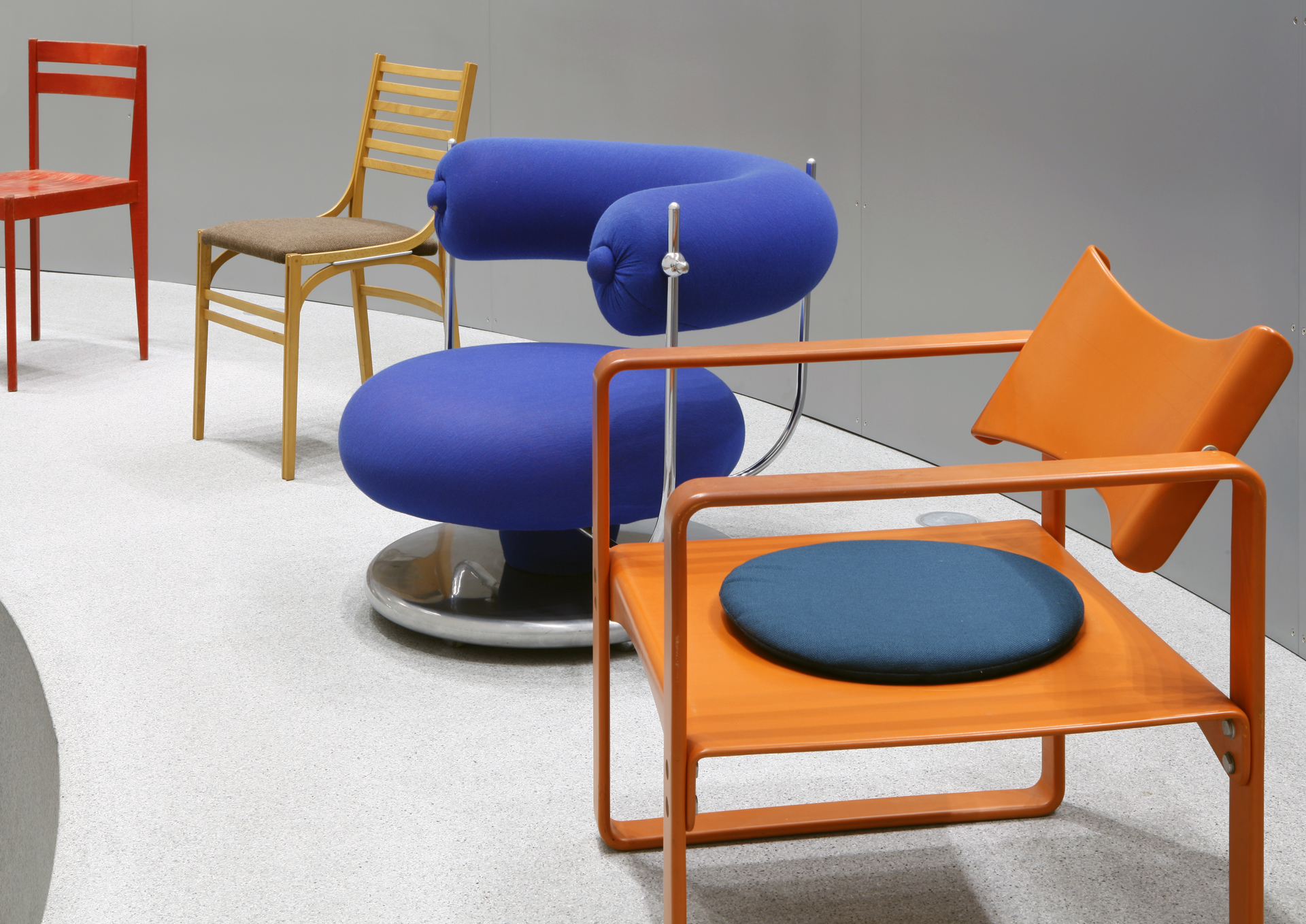 View of the Thonet & Design exhibition. Close-up of four chairs lined up one behind the other. A view of the Thonet & Design exhibition. Close-up of four chairs lined up one behind the other. The first one is made of wood, followed by a blue, round armchair and two more wooden chairs in beige and red.