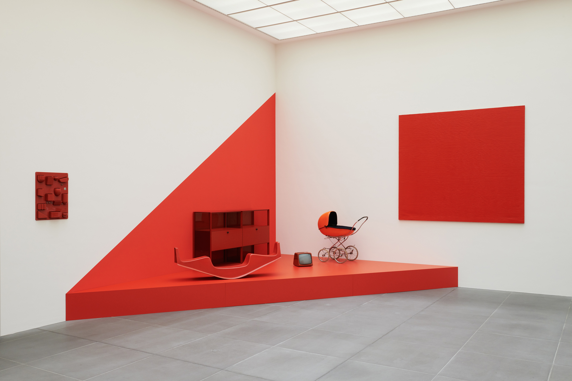 Exhibition view. Four objects in red colour (pram, television, rocker, shelf) can be seen on a red-painted pedestal with a triangular floor plan. On the walls (left of the pedestal) the red wall container ‘Utensilo’ and a painting in red colour (right).