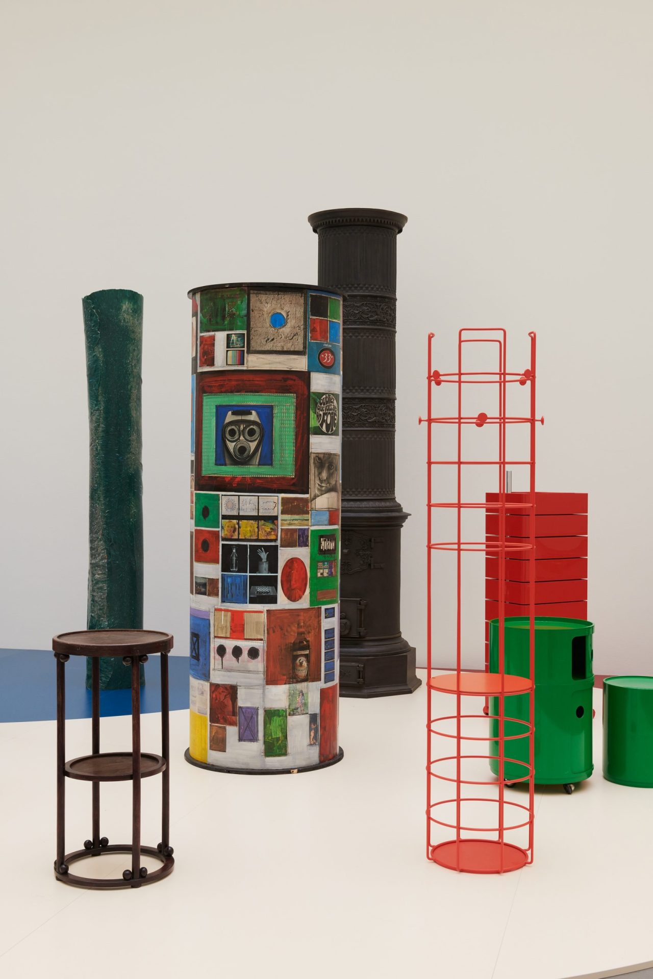 View of the exhibition. Objects on a light-coloured pedestal with circular floor plans (green container, red metal shelf, brown wooden side table, behind it an ‘advertising pillar’ (a pillar placed on the pavement of streets to which posters are stuck) and a cast iron stove.