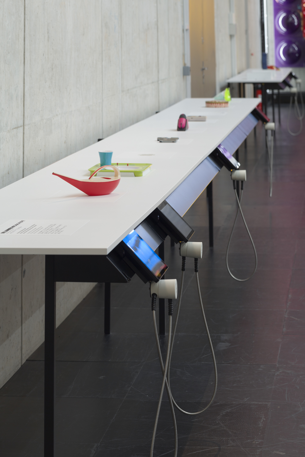 Picture in portrait format, view of the two touch tables with screens on a black rail. The table top is white. Several colourful tactile objects are attached. A watering can can be seen at the front.