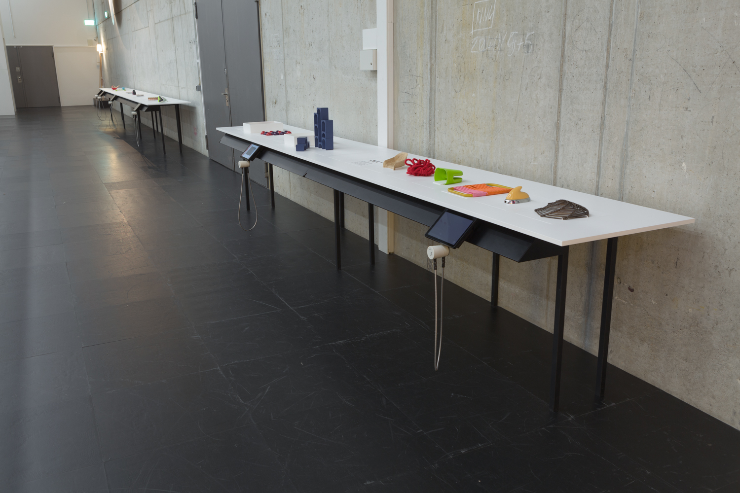 The picture shows the touch station in the exhibition space: a table stands in front of a grey concrete wall. It has a thin, black table frame and a white table top. There are several colourful tactile objects on it.