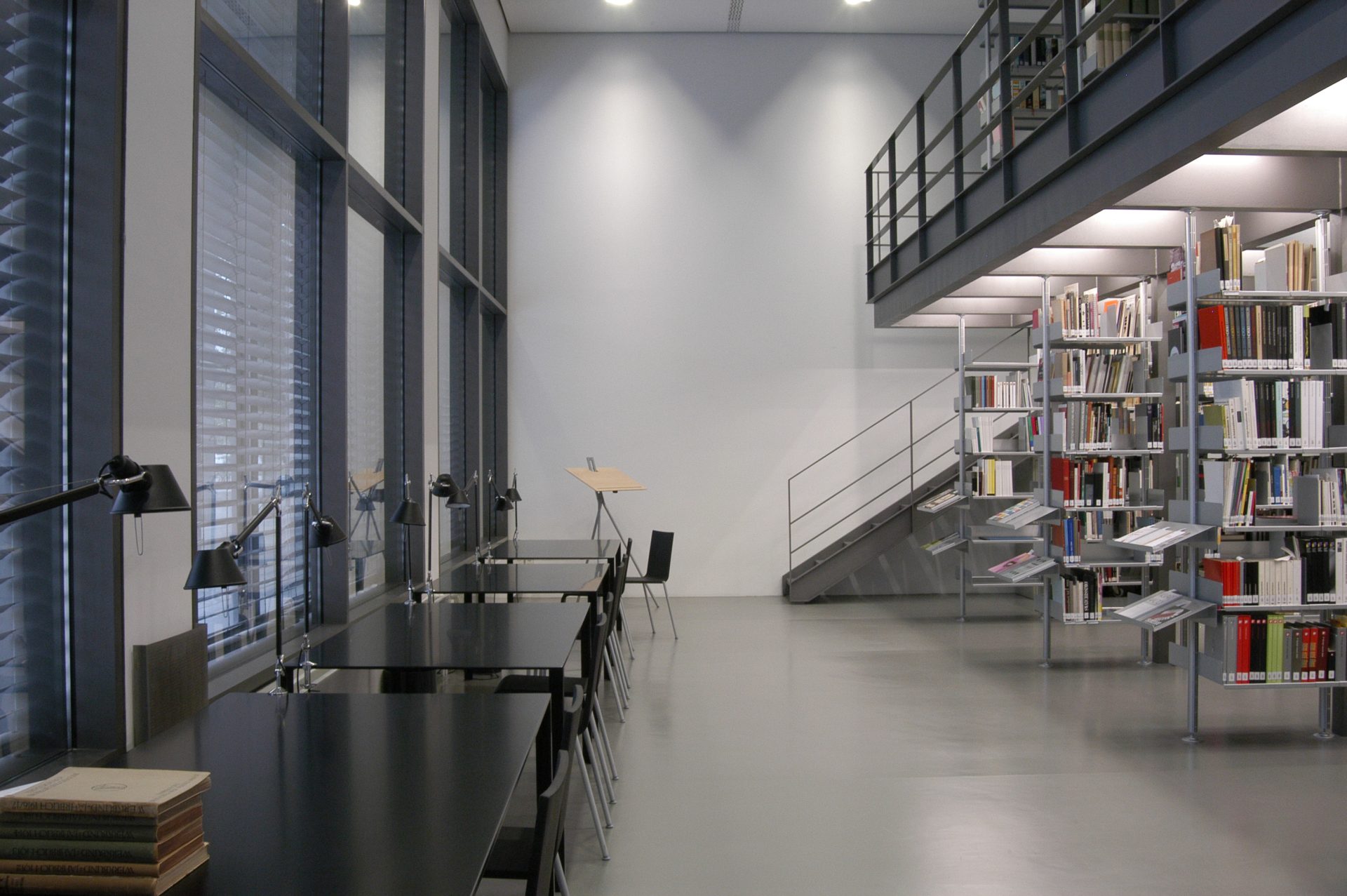 Zu sehen ist ein Blick in die helle Bibliothek. Der Boden ist grau, die Wände sind weiß. Links sind an einer großen Fensterfront schwarze Tische und Stühle. Rechts sind offene, weiße Regale. Man blickt auf eine Treppe, die auf die Empore links führt. 