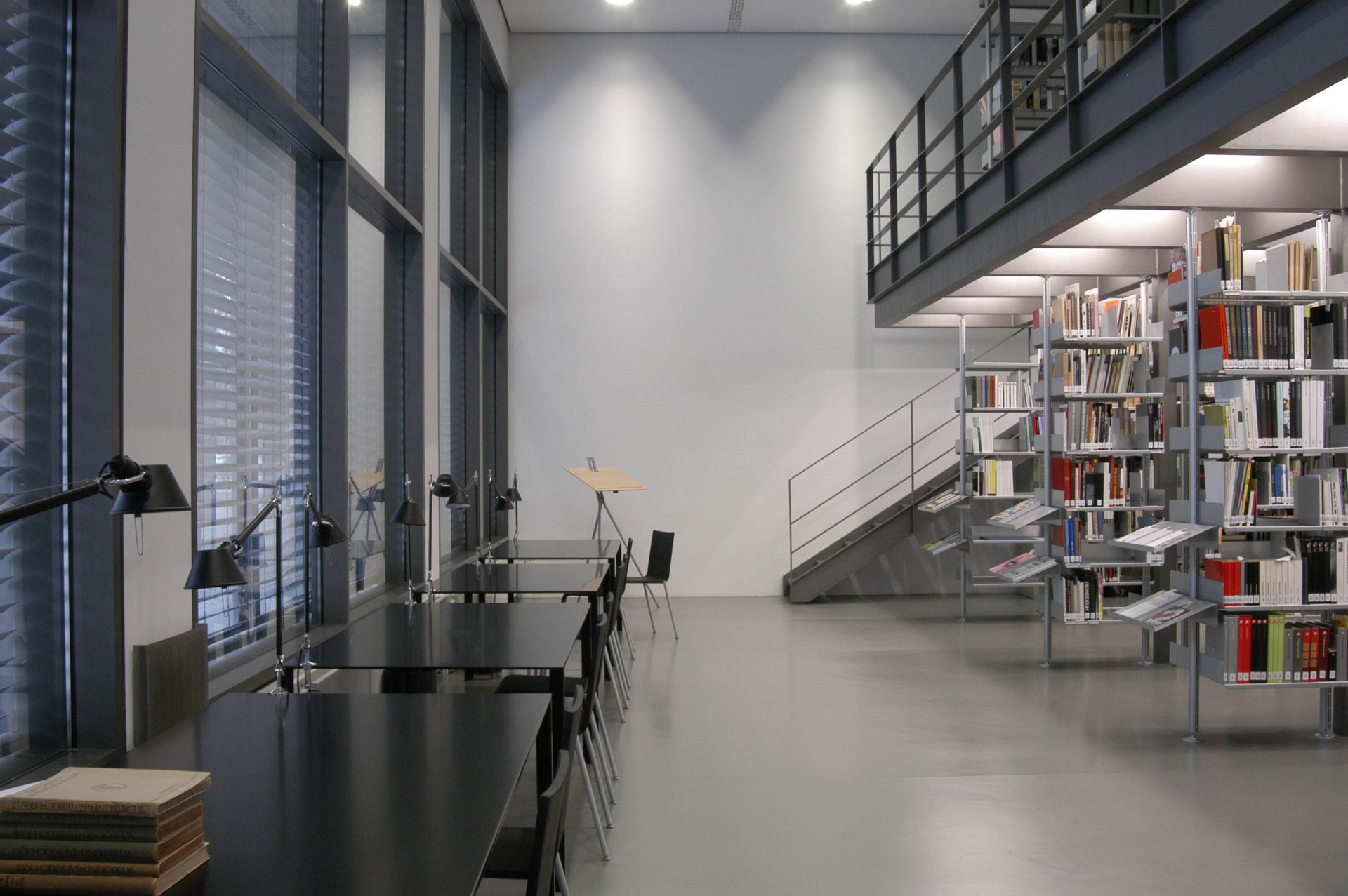 Zu sehen ist ein Blick in die helle Bibliothek. Der Boden ist grau, die Wände sind weiß. Links sind an einer großen Fensterfront schwarze Tische und Stühle. Rechts sind offene, weiße Regale. Man blickt auf eine Treppe, die auf die Empore links führt.