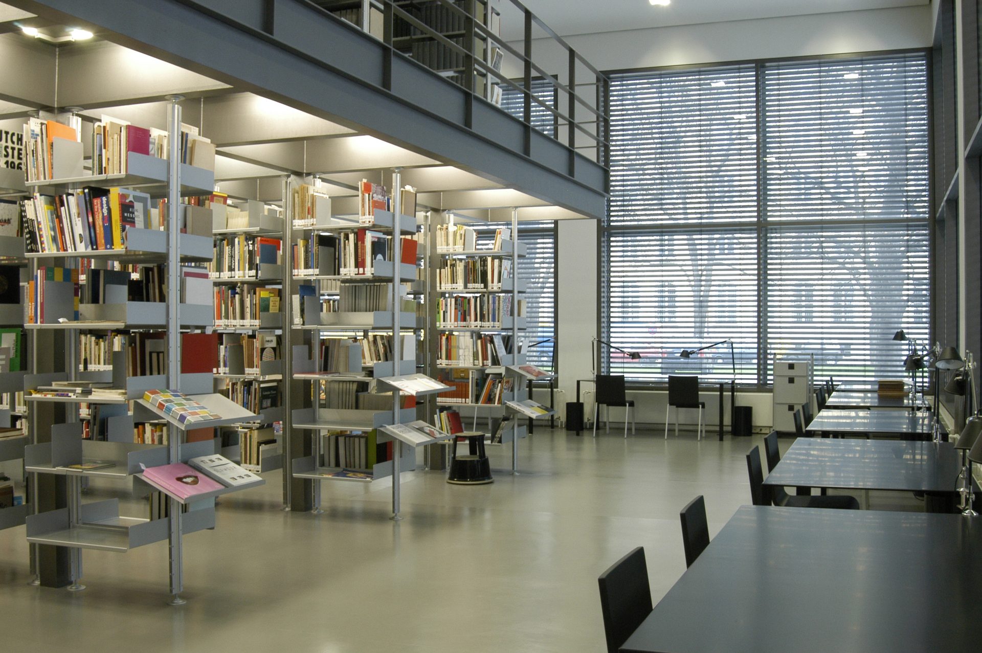 Das Bild zeigt die Bibliothek mit hohen Decken, Glasfenstern und Jalousien. Links sind gefüllte Bücherregale.