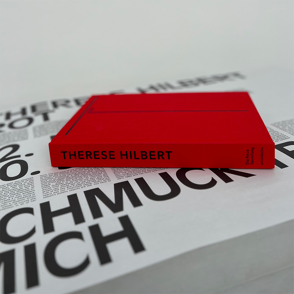 Exhibition Catalogue on white board with black lettering. The book is red and it says Therese Hilbert.