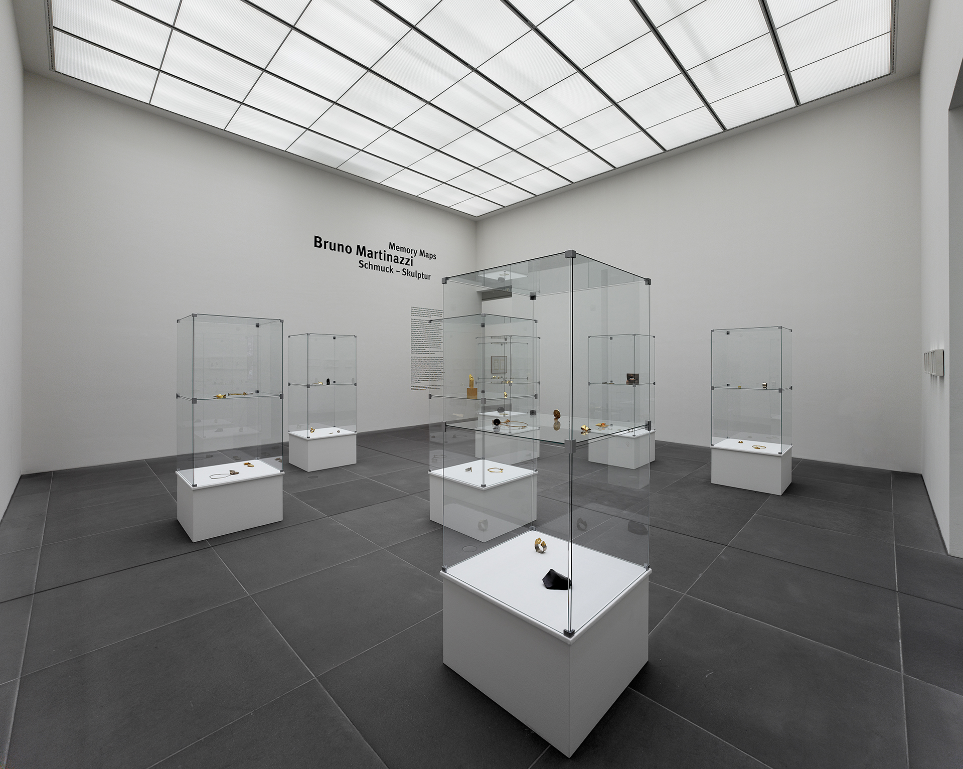 View of the exhibition. You can see a light-flooded exhibition room with a large ceiling window. Spread around the room are eight tall glass display cases with white plinths. Individual pieces of jewelry can be seen in them.