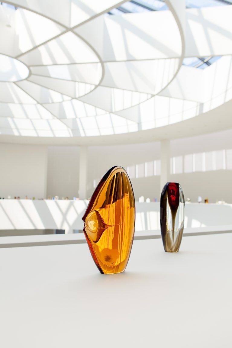 View of the museum with roof construction in the background. You can see two glass sculptures in orange and black-transparent.