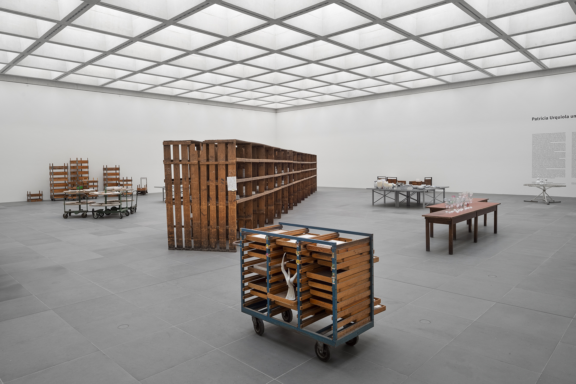 Large, bright exhibition room. In the foreground, a wooden trolley with a three-armed white sculpture. There is also a large wooden shelf and several tables with porcelain tableware and glasses.