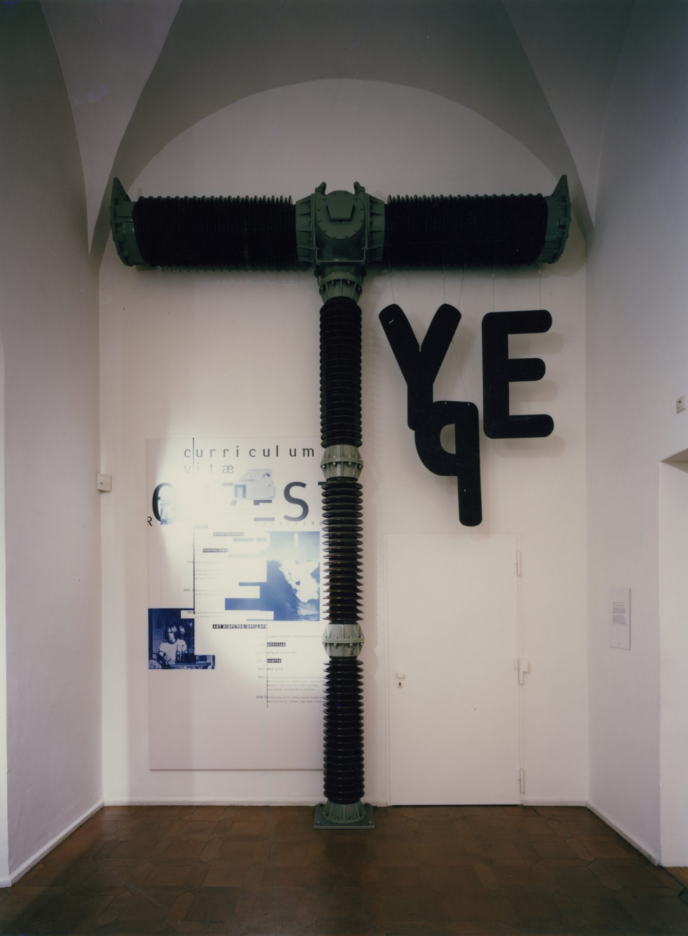 Ein großes Objekt in einem weißen schmalen Raum mit Gewölbe. Es erinnert von den Form her an einen Presslufthammer, an ihm hängen rechts überdimensional groß die Buchstaben "Y", "P" und "E". Links ein Plakat.