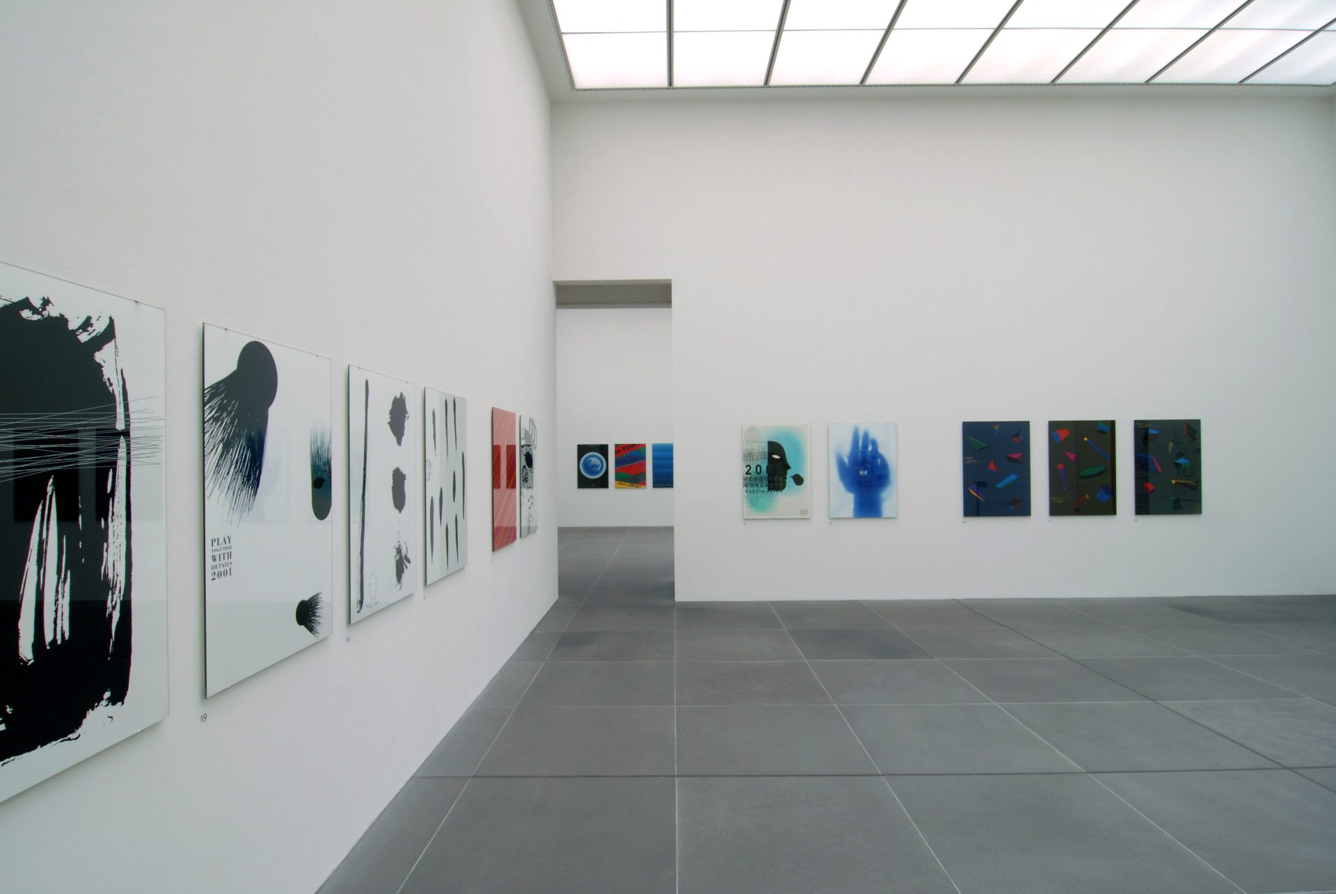 Zu sehen ist ein Ausstellungsraum des Museums, ein sogenannter White Cube, in dem an den Wänden verschiedene farbige wie schwarz-weiß Plakate des Künstlers hängen. Sie sind horizontal aneinander gereiht und ungefähr auf Augenhöhe angebracht.