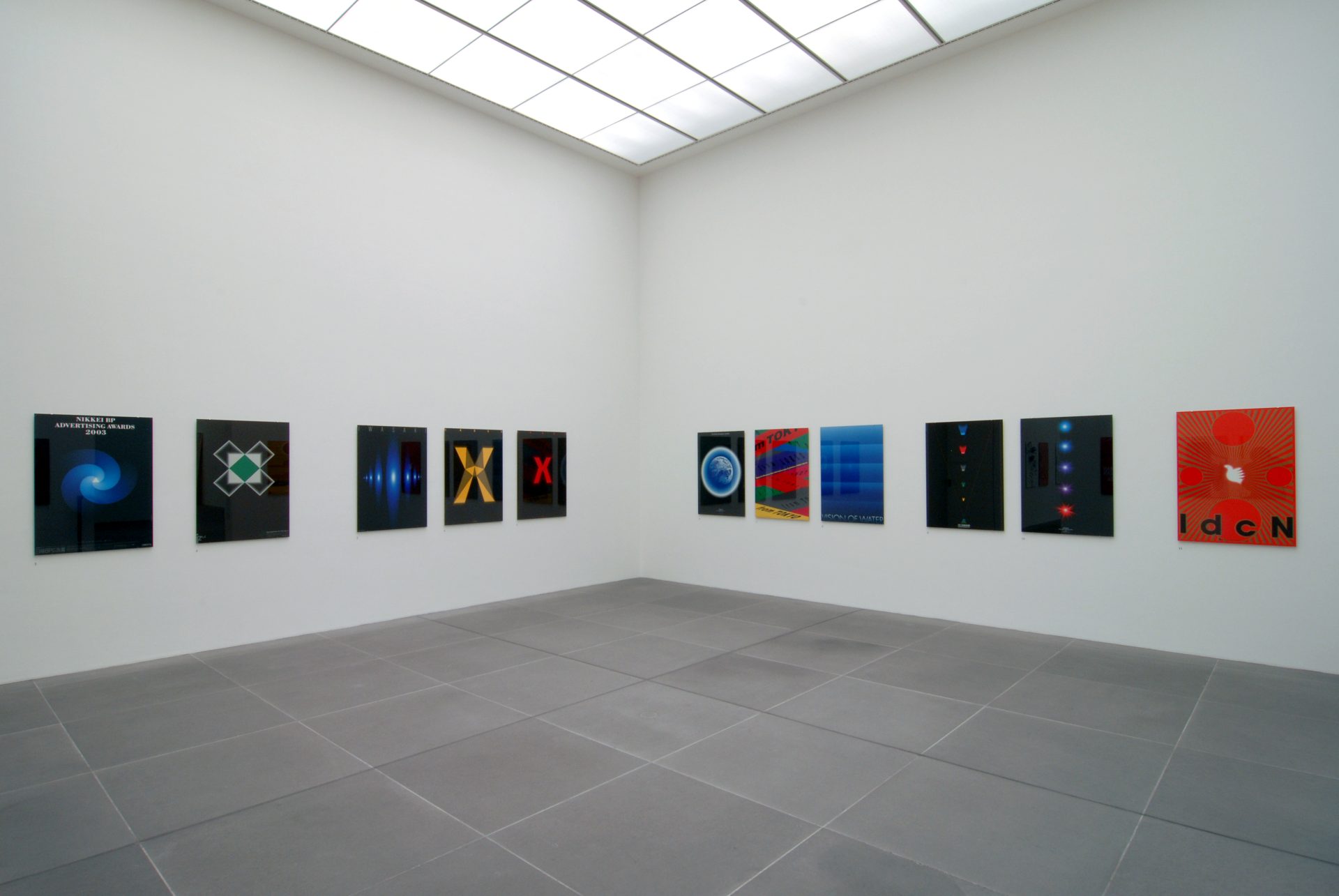 Zu sehen ist ein Ausstellungsraum des Museums, ein sogenannter White Cube, in dem an den Wänden verschiedene farbige Plakate des Künstlers hängen. Sie sind horizontal aneinander gereiht und ungefähr auf Augenhöhe angebracht.