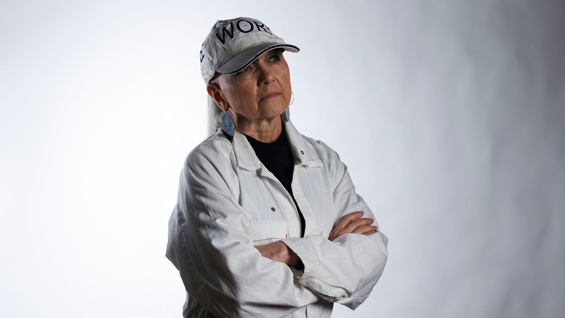 Portraitfoto Susan Cohn. Sie ist eine weiße Frau über 50 Jahre. Sie trägt eine weiße Jeans-Bluse und ein weißes Cappy mit der schwarzen Aufschrift Words. Sie ist im Seitenprofil und hat die Arme verschränkt.