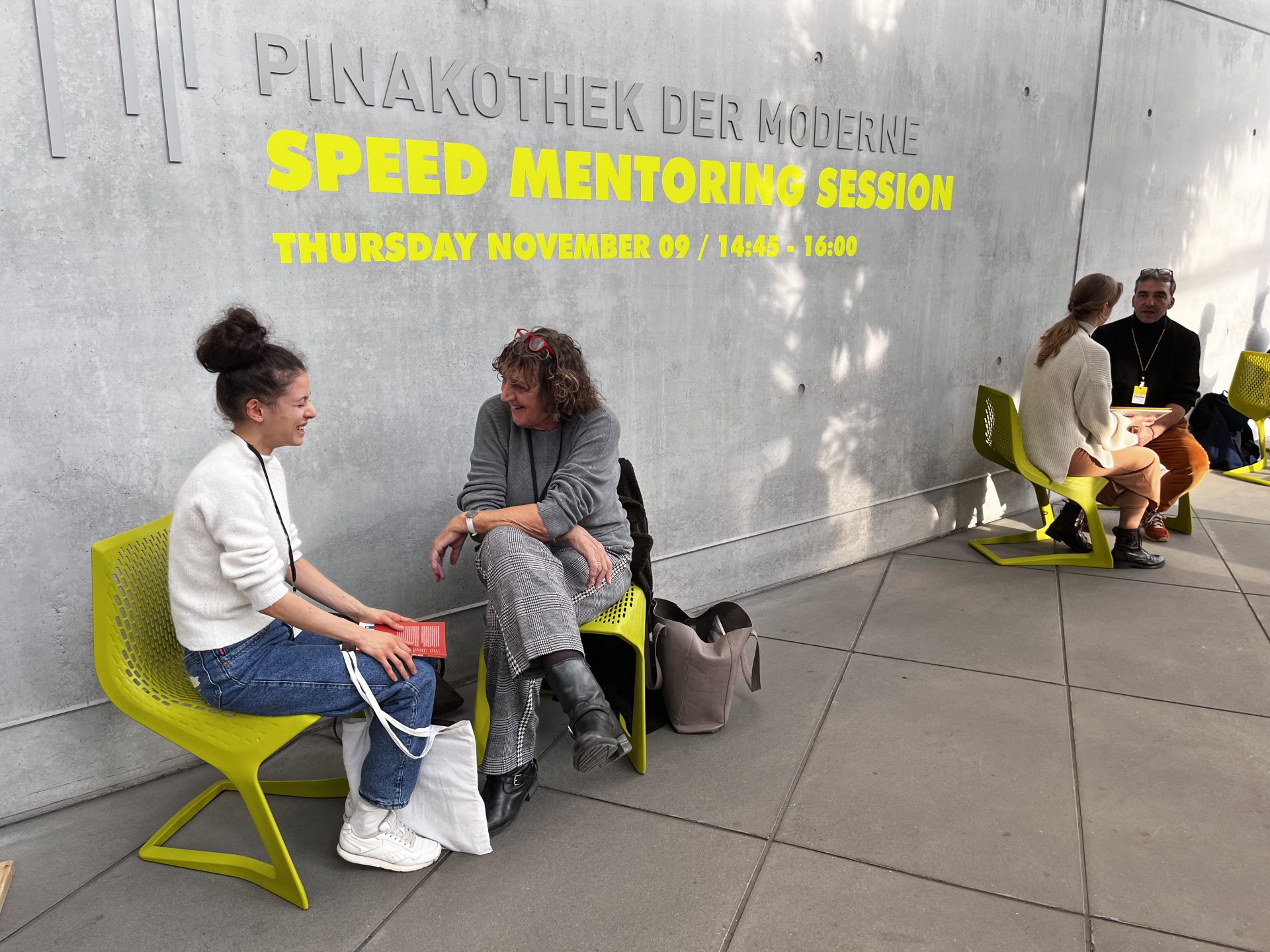 Zwei Teilnehmerinnen unterhalten sich auf der Terrasse der Pinakothek der Moderne. Sie sitzen auf hellgrünen Stühlen. Auf der Betonwand im Hintergrund steht in der Schriftart der Veranstaltung "Speed Monitoring Session" unter der Aufschrift Pinakothek der Moderne. Rechts sitzen zwei weitere Personen an der Wand.