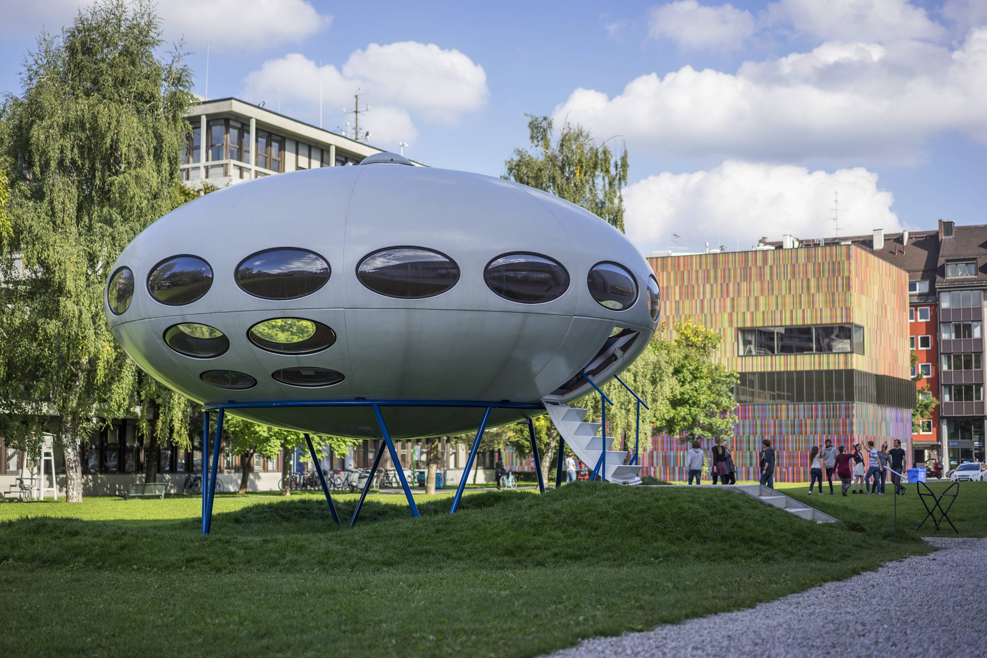 The picture shows the mobile home Futuro, which resembles a UFO. It stands on a meadow in front of the Pinakothek der Poderne. In the background you can see the square, colorful building of the Museum Brandhorst.