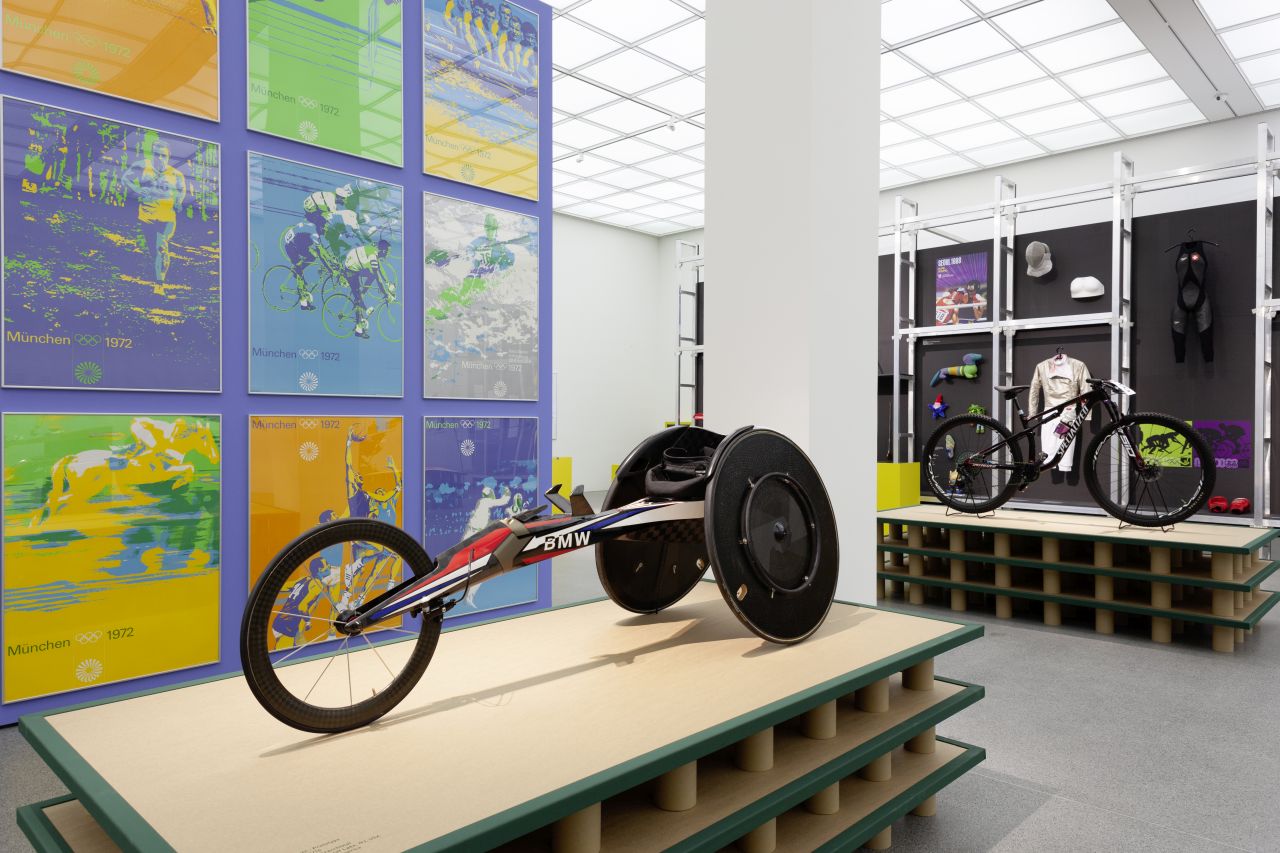 In the foreground BMW racing wheelchair, behind it sports posters from Team Otl Aicher. In the background, Specialized mountain bike in front of a black wall of shelves with mascots, timing devices and sports equipment from different eras