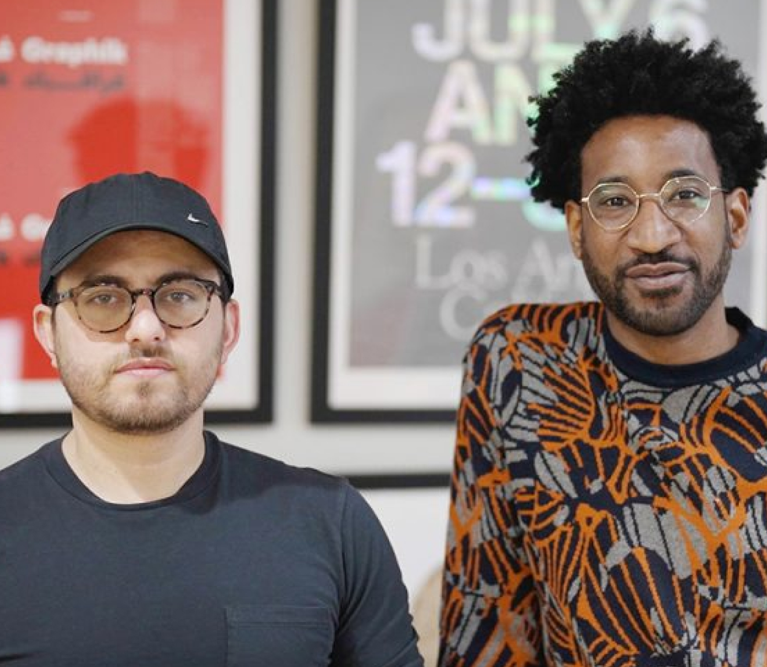 Two men look into the camera. The one on the right is a white man in his mid-30s with round horn-rimmed glasses and a black cap. He wears a 3-day beard and a black T-shirt. The second has metal glasses, also a 3-day beard and is a Black Person of Colour. He is wearing an orange and black patterned jumper.