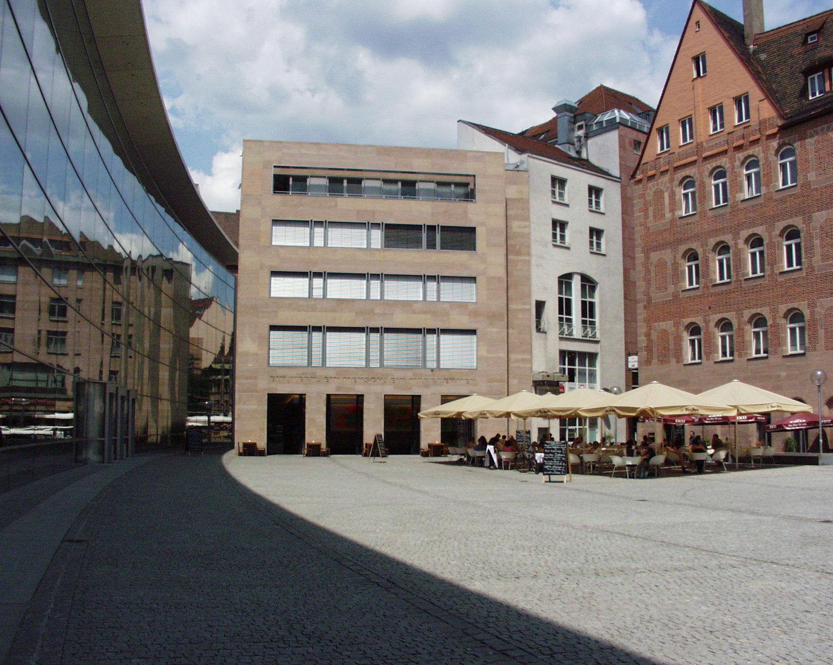 Außenansicht Neues Museum Nürnberg bei Tag: Klarissenplatz mit Backsteingebäude, links Glasfront des Museums, rechts weitere Gebäude und Sonnenschirme