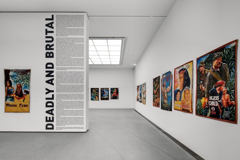 Blick in die Ausstellung. Zu sehen sind mehrere großformatige Filmplakate an den Wänden. Der Raum wird durch eine Zwischenwand geteilt. Im vorderen Teil ist der Schriftzug "Deadly and Brutal" in Versalien vertikal an die Wand aufgebracht. Direkt rechts daneben zieht sich der Ausstellungstext über die Wand - von der Decke bis zum Boden. Im hinteren Teil ist ein großes Deckenfenster zu sehen, durch welches der Raum erhellt wird. Auf den handgemalten Filmplakaten an den Wänden sind in knalligen Farben Figuren/Personen zu sehen.