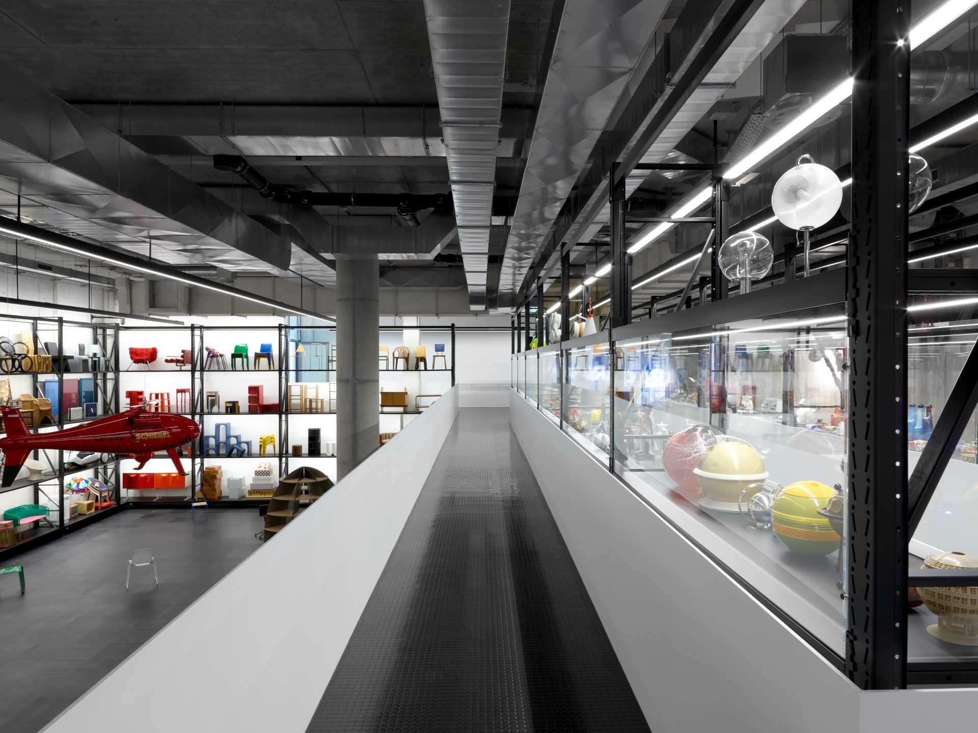 The picture shows a corridor with a black floor and white ballustrades. On the right are glass cabinets, on the left you can see into the X-D-E-P-O-T with high shelves. The shelves contain various colourful collection objects, such as furniture.