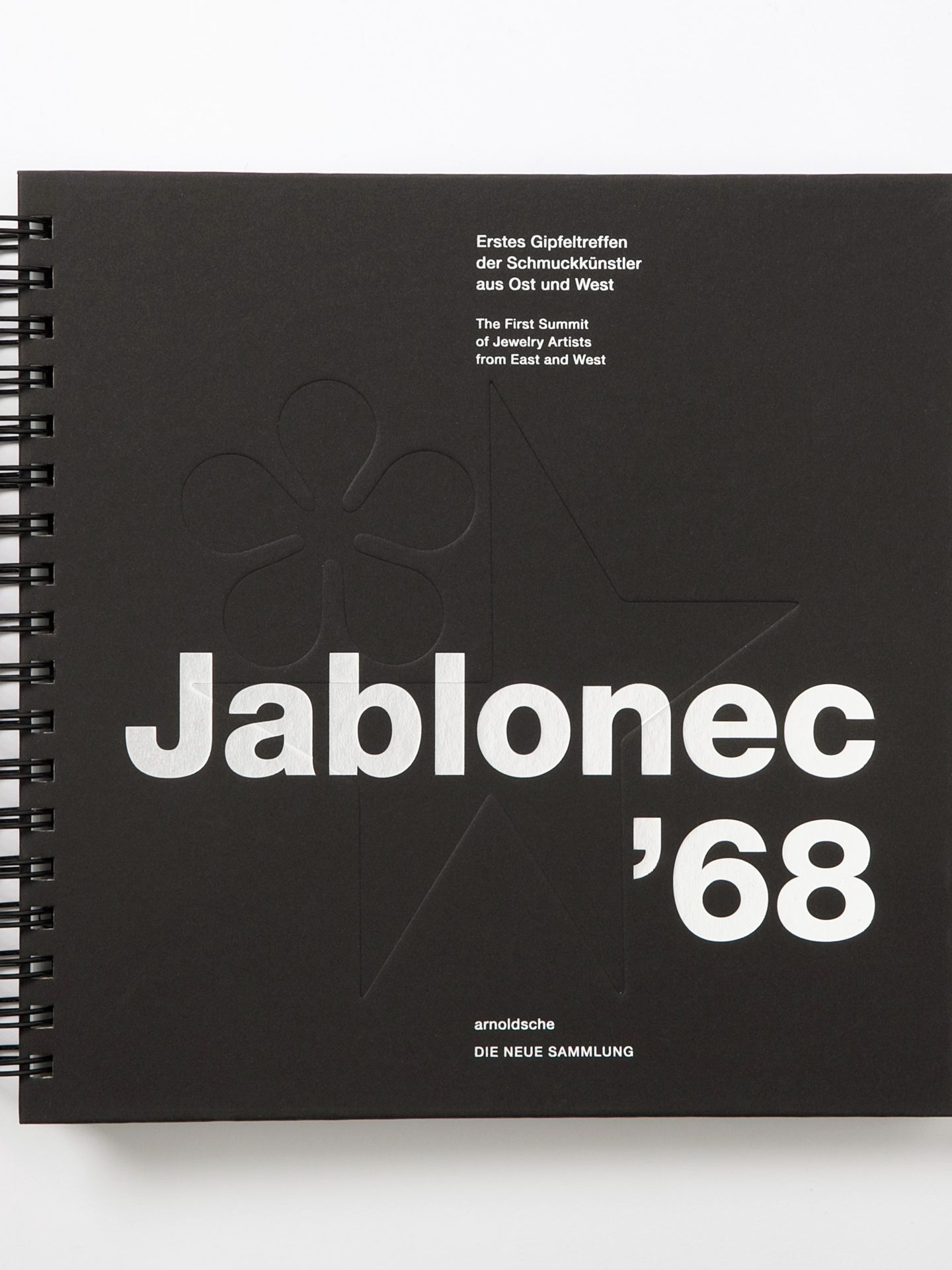 Schwarzer Einband eines Ausstellungskatalogs mit Prägung in Blumenform und Stern. Ringbindung und weiße Beschriftung.
