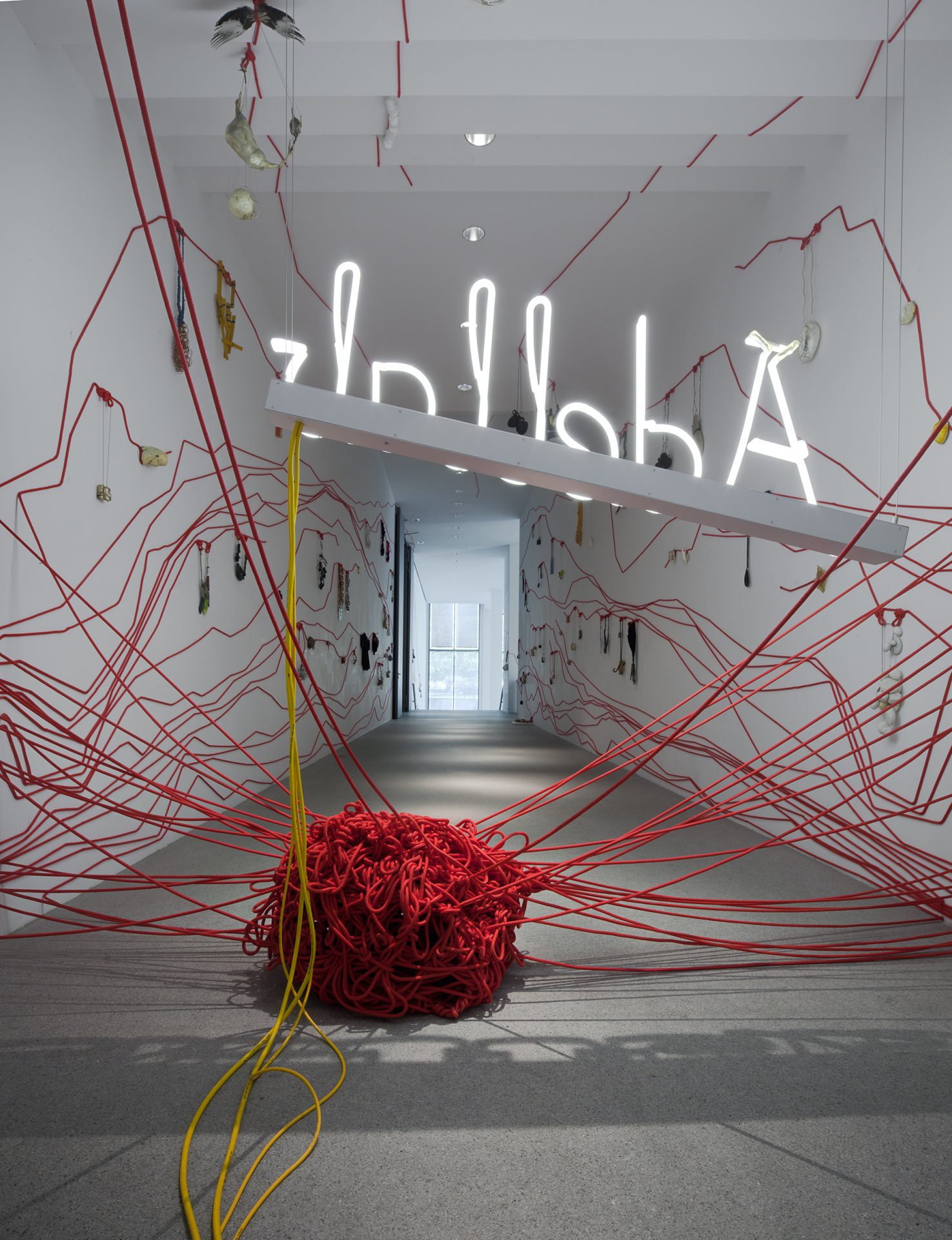 View of the exhibition. You can see a large red ball of wool lying on the floor in the middle of a corridor. The word Ädellab is written in neon letters above the ball of wool. Countless threads extend from the ball of wool, running along the walls to the right and left and across the ceiling. The individual pieces of jewelry in the exhibition are attached to the threads.