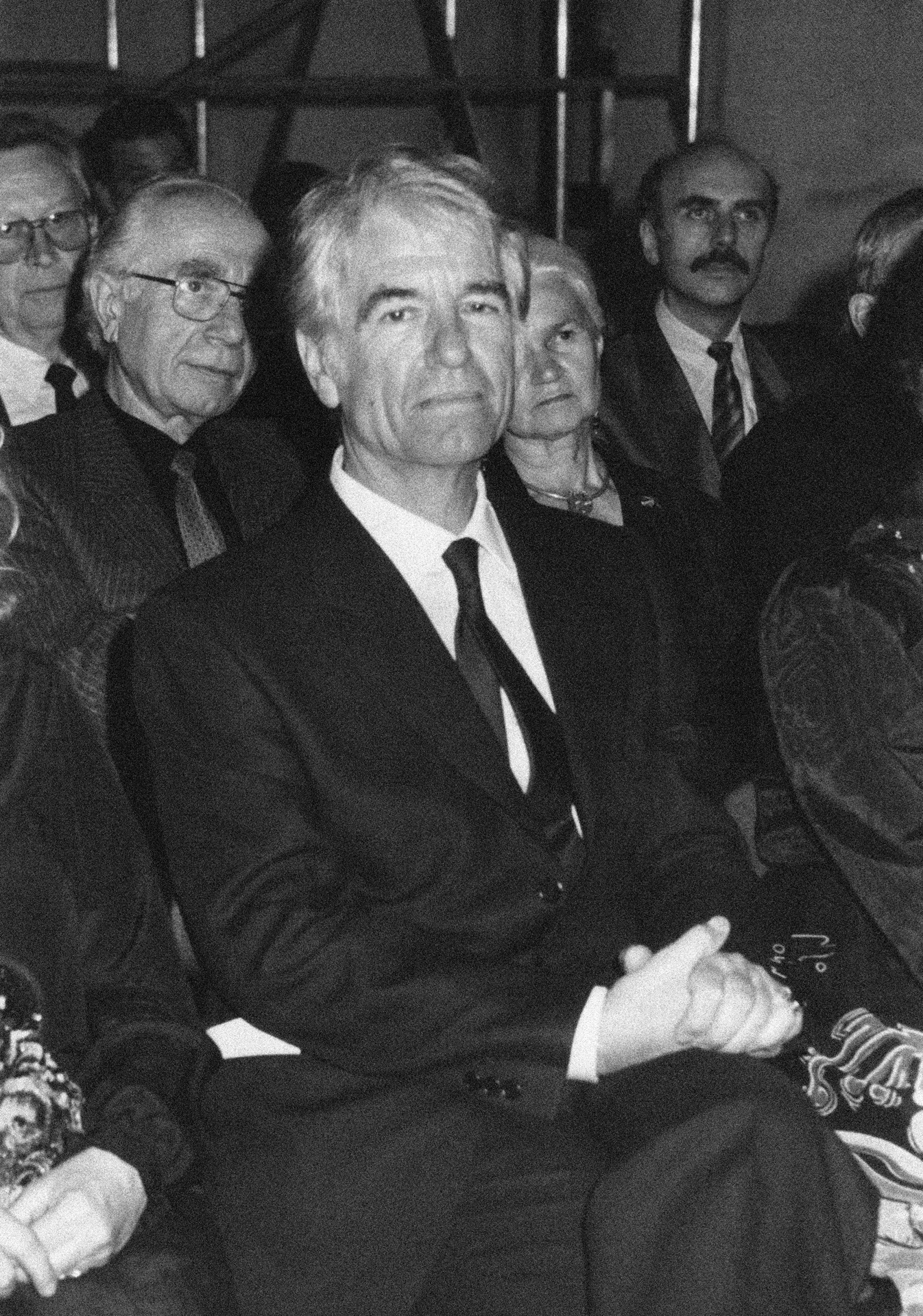 Black and white portrait photo. A man in his 70s is sitting on a chair in the audience, with other people sitting behind him. The man is wearing a black suit, a tie and has his hands folded in his lap. His face is long, his thick hair is white, his eyebrows are prominent. He has no beard.