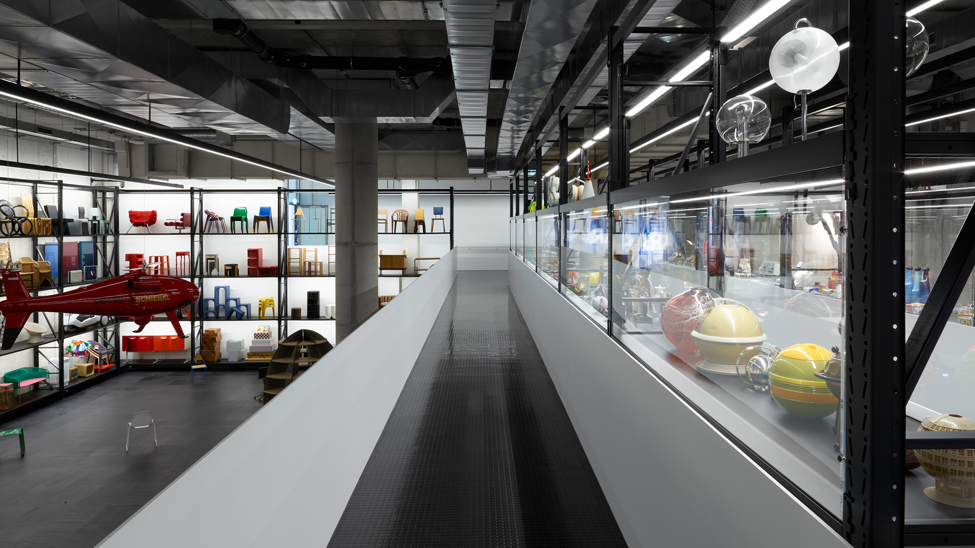 Room view of the permanent exhibition. You can see an industrial cellar room with a long corridor in the middle. To the left and right, individual exhibits from the museum's collection are stored on ceiling-high shelves and in glass cabinets. These include chairs in various colors and shapes, a red helicopter hanging from the ceiling and various designer lamps.