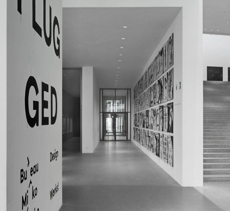 View into the exhibition. You can see a corridor with exhibits (posters) in black and white placed above and next to each other on the right-hand wall.