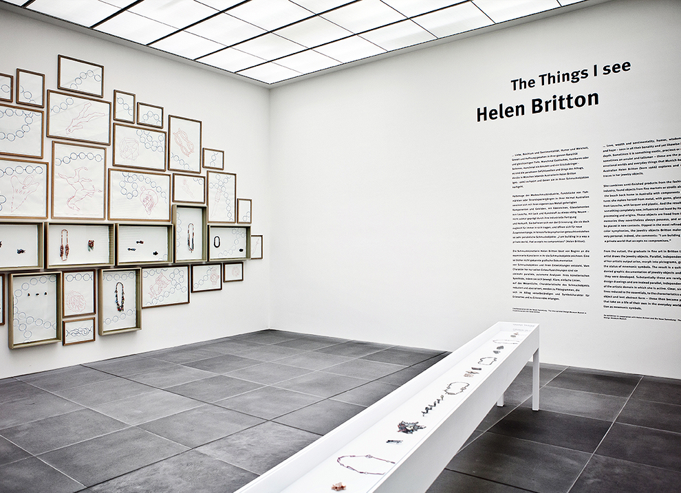The picture on the left shows several framed drawings hanging close together on the wall. On the right is the wall text for the exhibition in black on white. Below it is a white display case with pieces of jewellery.
