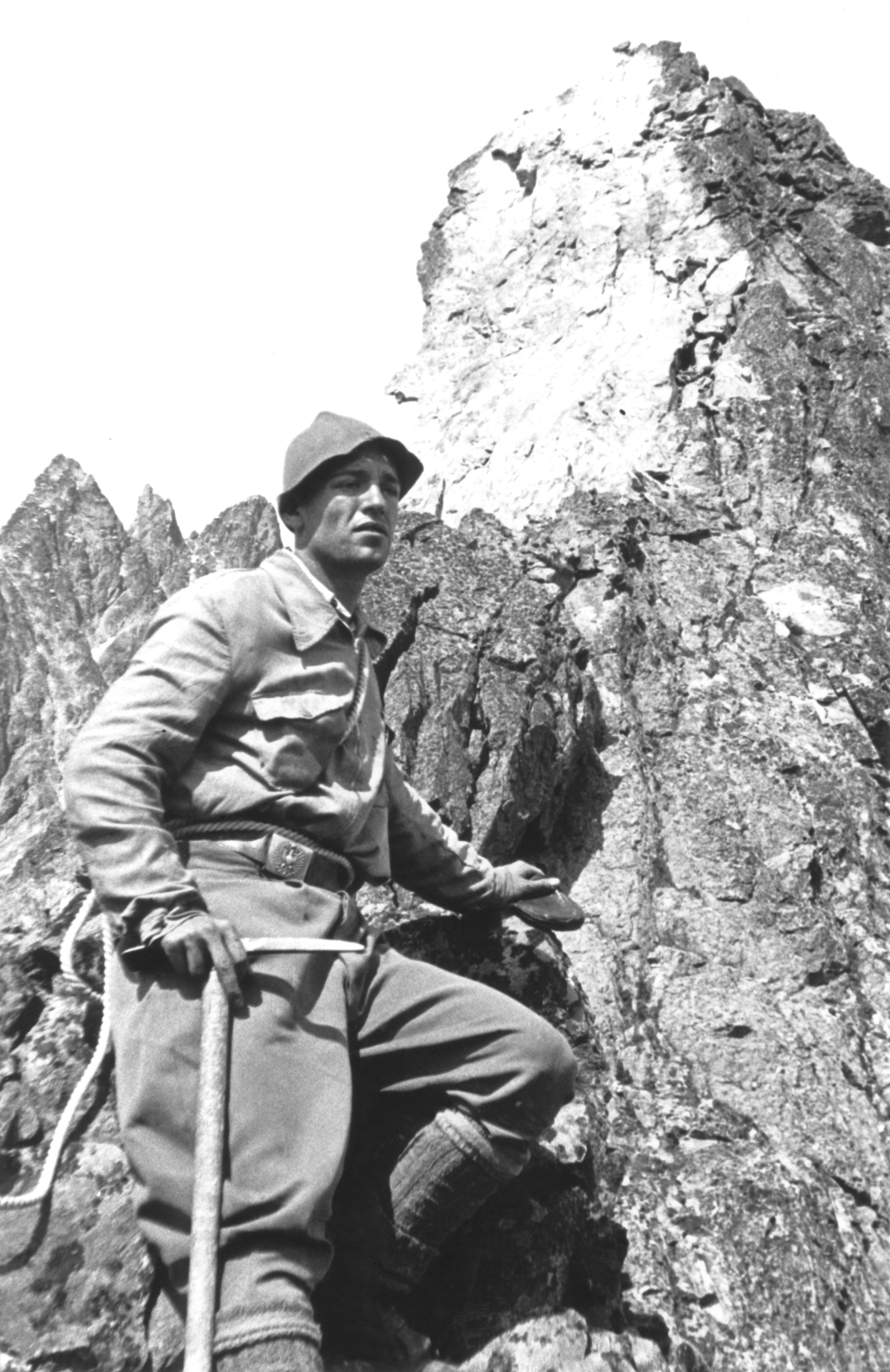 On display is a black and white photograph showing Bruno Martinazzi climbing a mountain peak. The artist is wearing a hat and linen clothes and is holding a chisel in his hand.