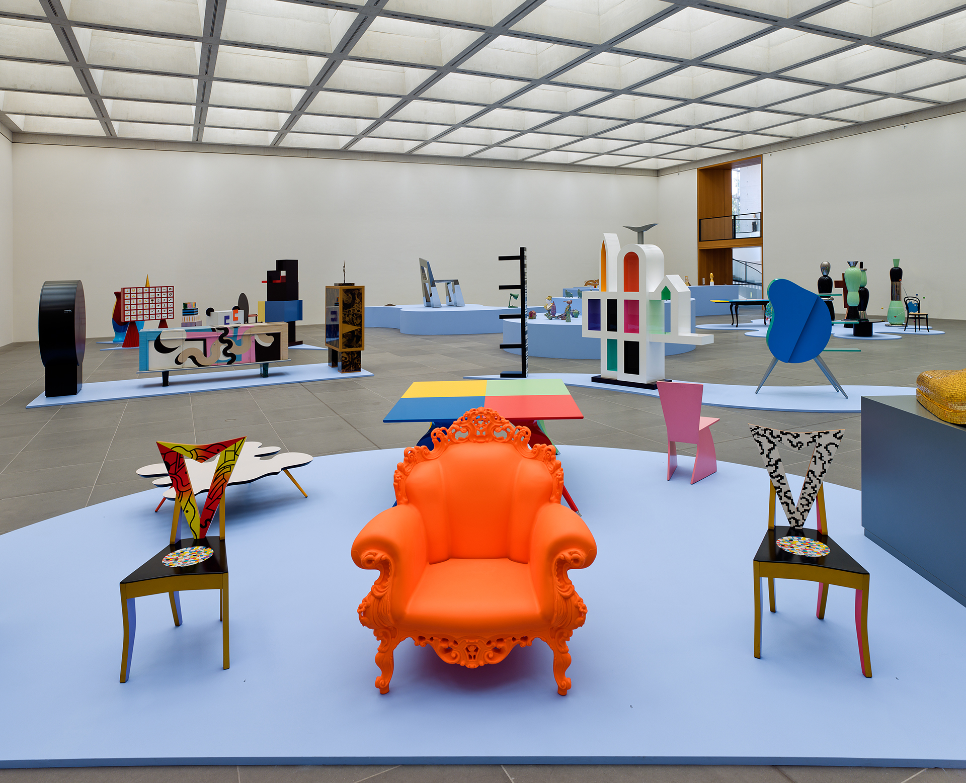 View of the exhibition. On display are several design objects in various bright colors and shapes. In the foreground are three chairs - on the left and right a wooden chair with a triangular backrest and in the middle an orange armchair with ornaments. The group of chairs is placed on a light blue, round background, as are the groupings of individual exhibits in the background.