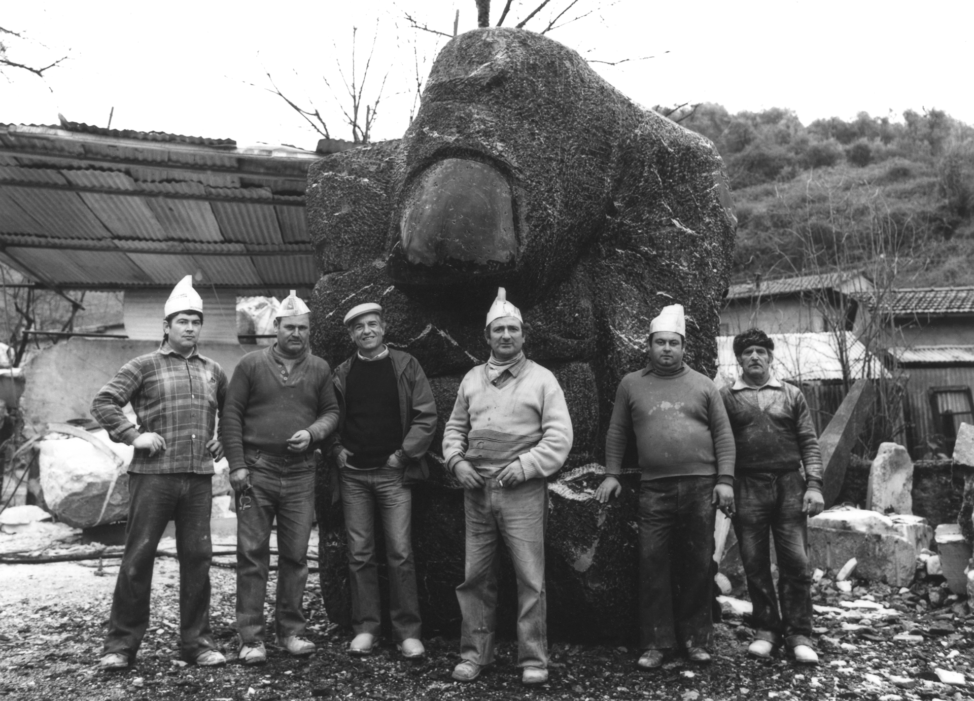 Schwarz-weißes Gruppenfoto. Sechs Männer mit Mützen stehen in Reihe vor einer Steinskulptur. Im Hintergrund sind zwei Häuser und ein Hügel.