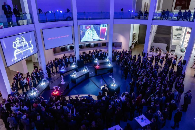 Foto into the Foyer of the Pinakothek der Moderne. Prof Sami Haddadin demonstrates the possibilities of collective machine learning at the Pinakothek der Moderne. You can see a lot of people and 6 tables with robotic arms. Above are Screens with a presentation.