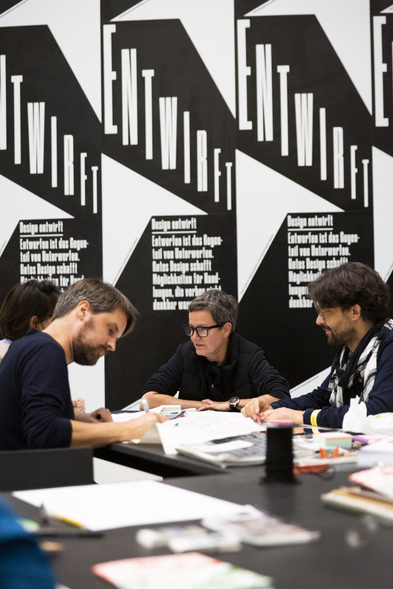 Fotografie eines Workshops. Vier Menschen sitzen an einem schwarzen Tisch. Ein Mann notiert etwas mit einem Stift. Im Hintergrund sind schwarz-weiße Plakate zu sehen.