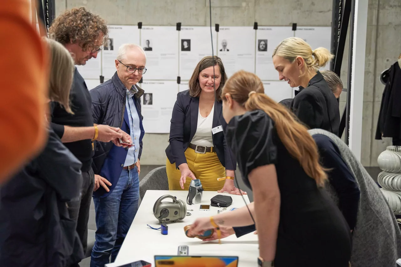 A group of people are standing around a table with various objects that they are touching. People are laughing. Posters from the workshop hang on a wall in the background.