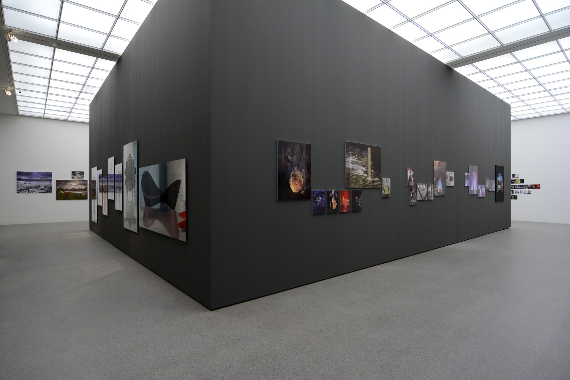 In a white exhibition room with an illuminated box ceiling, a black square of partition walls has moved in. On the walls there are high-contrast and high-quality photographs of design objects.