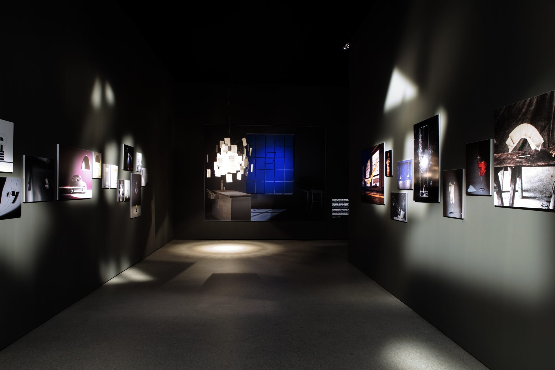 Blick in einen abgedunkelten Raum. Am Ende des Raums hängt eine Leuchte mit weiß-warmem Licht. Daneben scheint sich ein Fenster mit blauen Glasscheiben zu befinden. An den Wänden werden hochwertige Fotografien ausgestellt. Sie zeigen andere Leuchtobjekte.