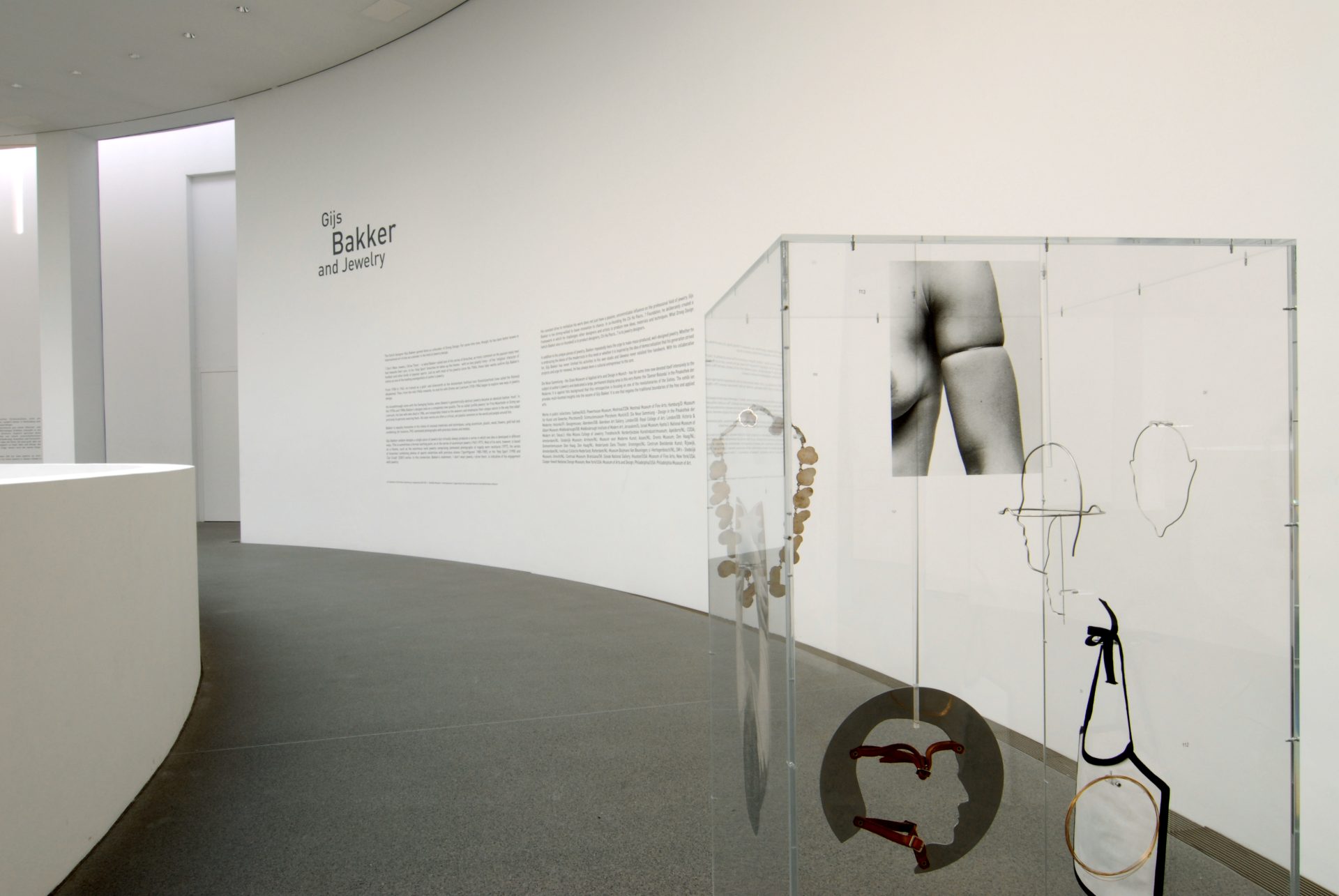 The picture shows the wall text of the exhibition. To the right in front of it is a tall glass display case with an installation of various floating metal objects and a black-and-white print of a body section. This one shows a bare shoulder with upper arm and chest. There is a bracelet on the upper arm.