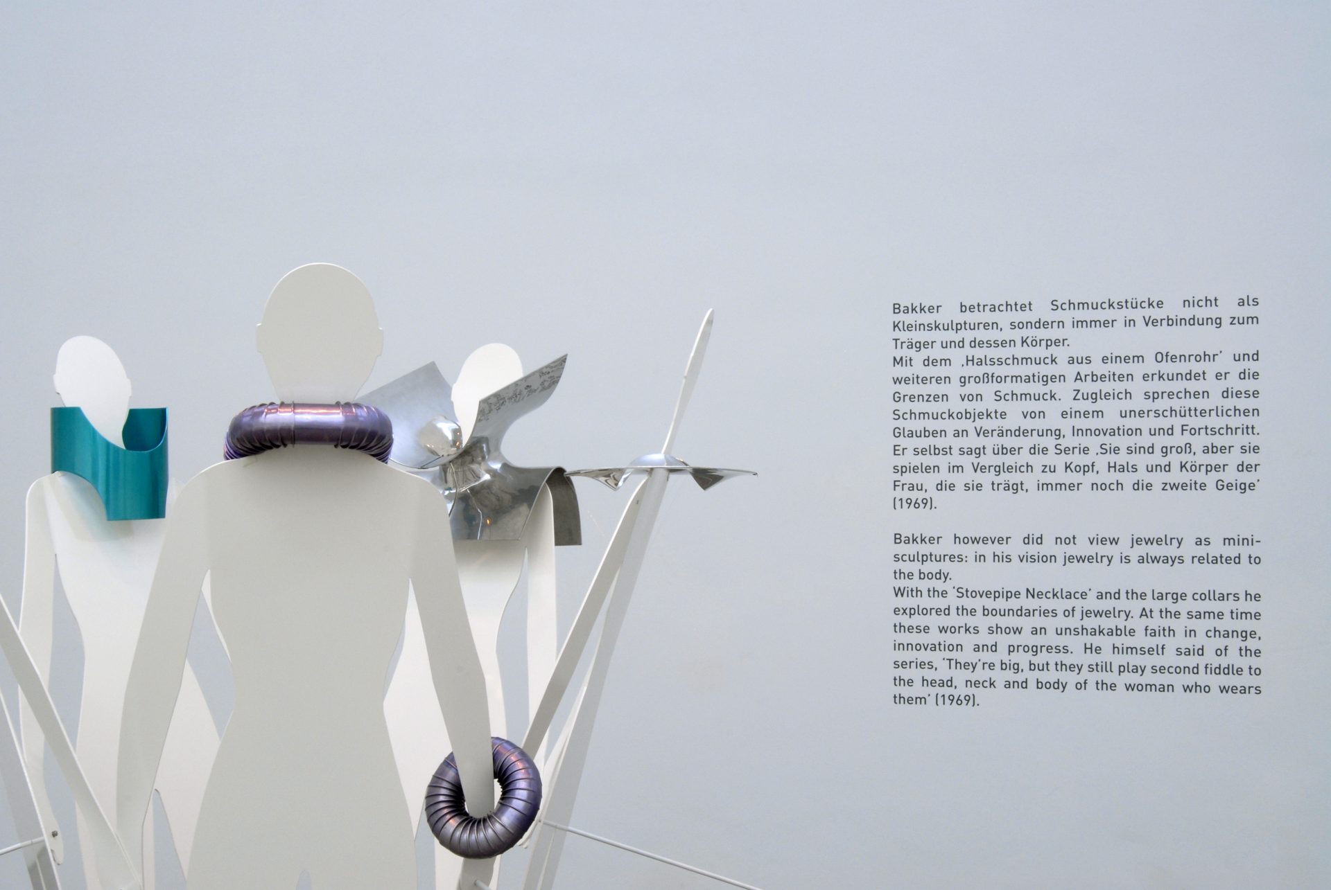 On the left are abstract metal figures wearing round decorative objects on their necks and arms. To the right is an informative wall text.