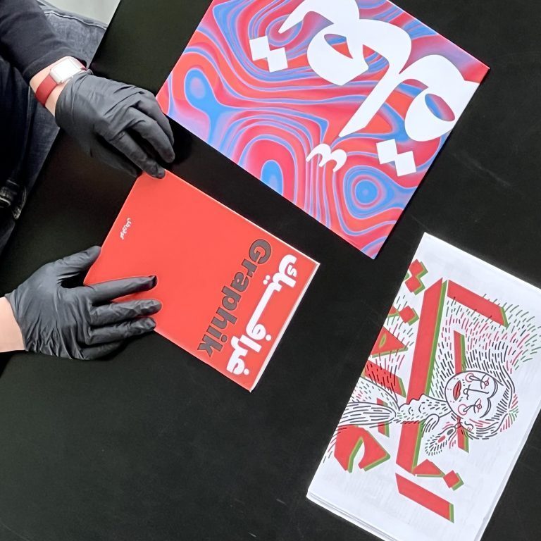 You can see several printed products lying on a table. Two hands with black rubber gloves are aligning one of the booklets. They are all red, white and blue with lettering.