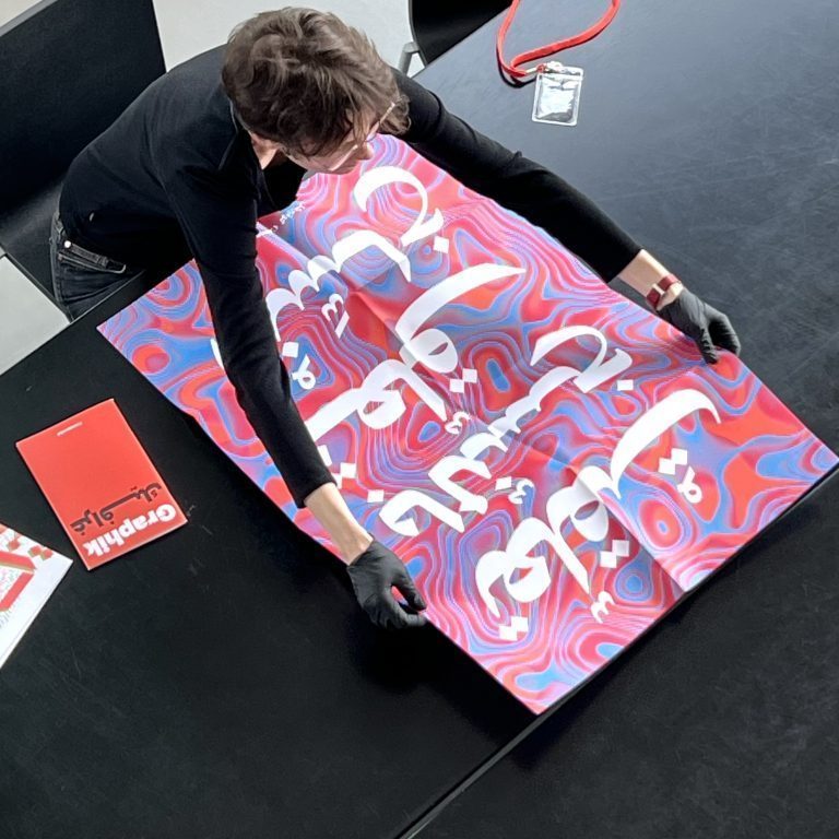 You can see a poster of a woman spread out on a black table. The poster has a white, Arabic font and an abstract background of blue, pink and red.