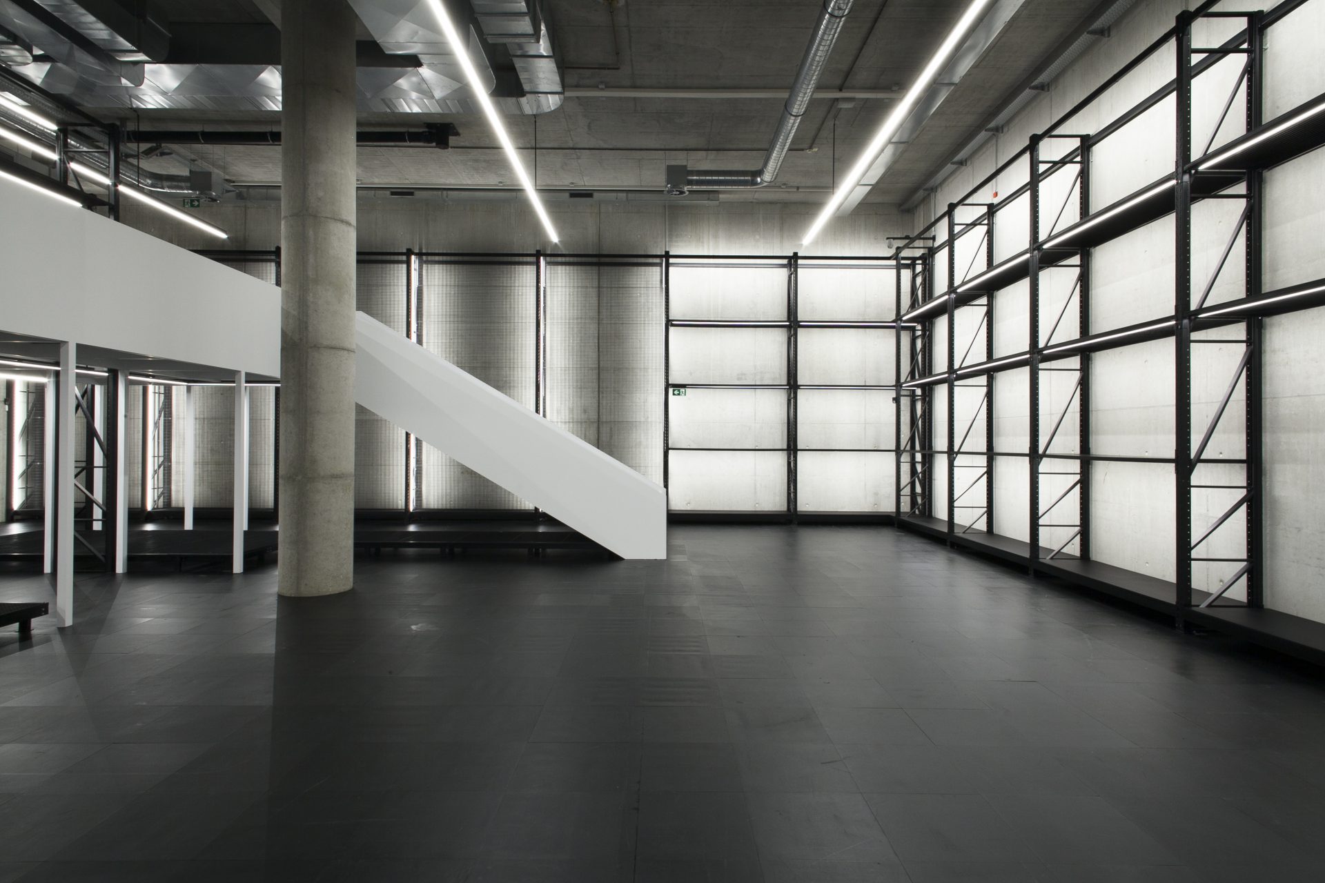 View of the empty showroom. The X-D-E-P-O-T is a large, high room with black shelves that reach up to the ceiling. The shelves are illuminated. The walls are made of grey concrete, the floor is black. On the left you can see a pillar and a white staircase coming from a walkway.
