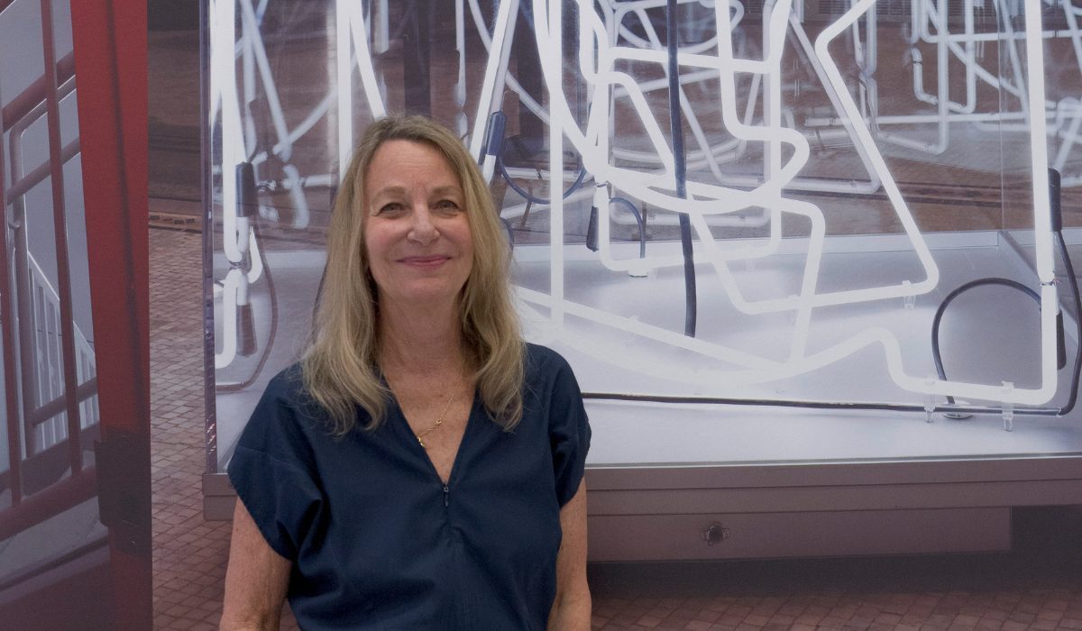 Man sieht Paula Scher vor einem ihrer Werke in der Ausstellung. Sie ist über 50, hat eine weiße Hautfarbe und schulterlange blonde Haare. Sie trägt ein blaues, kurzärmliges Oberteil.