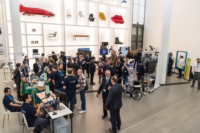 Photograph of an event in front of the type case. Various robotics research groups from TUM presented their work at the Pinakothek der Moderne. They sit at tables with their inventions. The event is well attended.