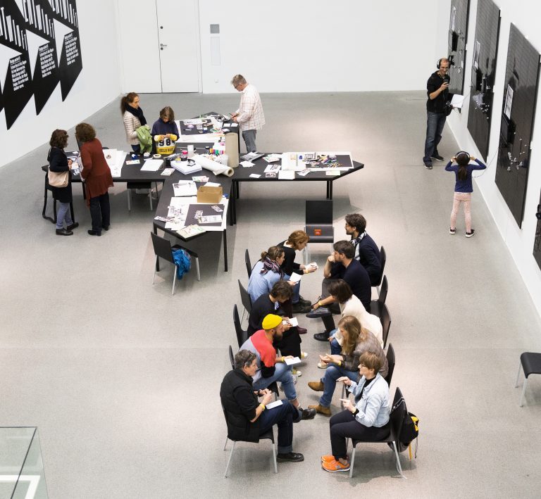 Foto eines Workshops. Menschen sitzen in einer Reihe und an einem schwarzen Tisch. Zwei weitere Personen stehen vor einer Wand. Alle scheinen damit beschäftigt zu sein, etwas zu schaffen und zu diskutieren.