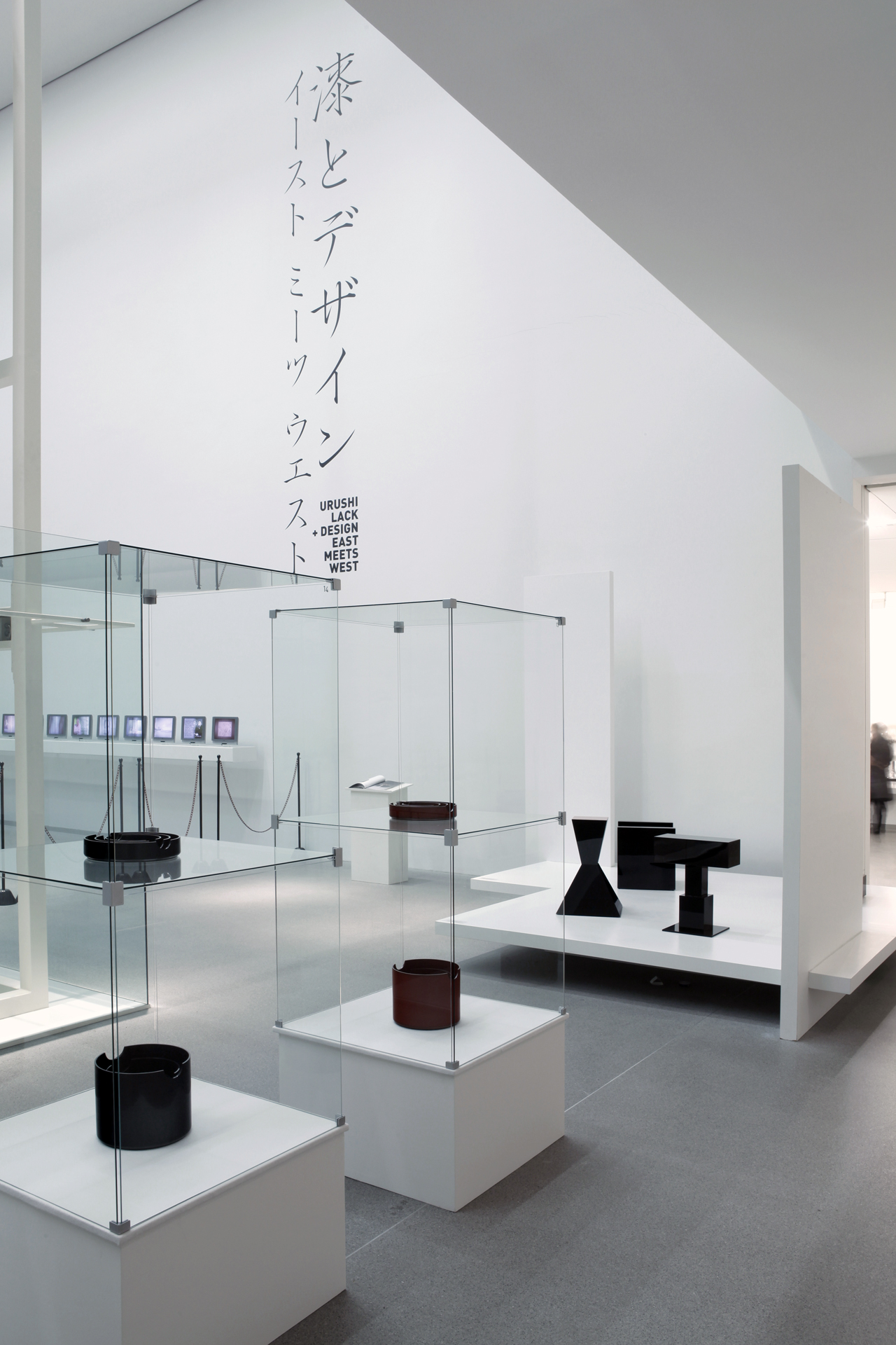 View of the exhibition. In a light-flooded room, you can see tall glass display cases and white pedestals in and on which black exhibits are placed. Japanese characters can be seen on the right-hand wall.