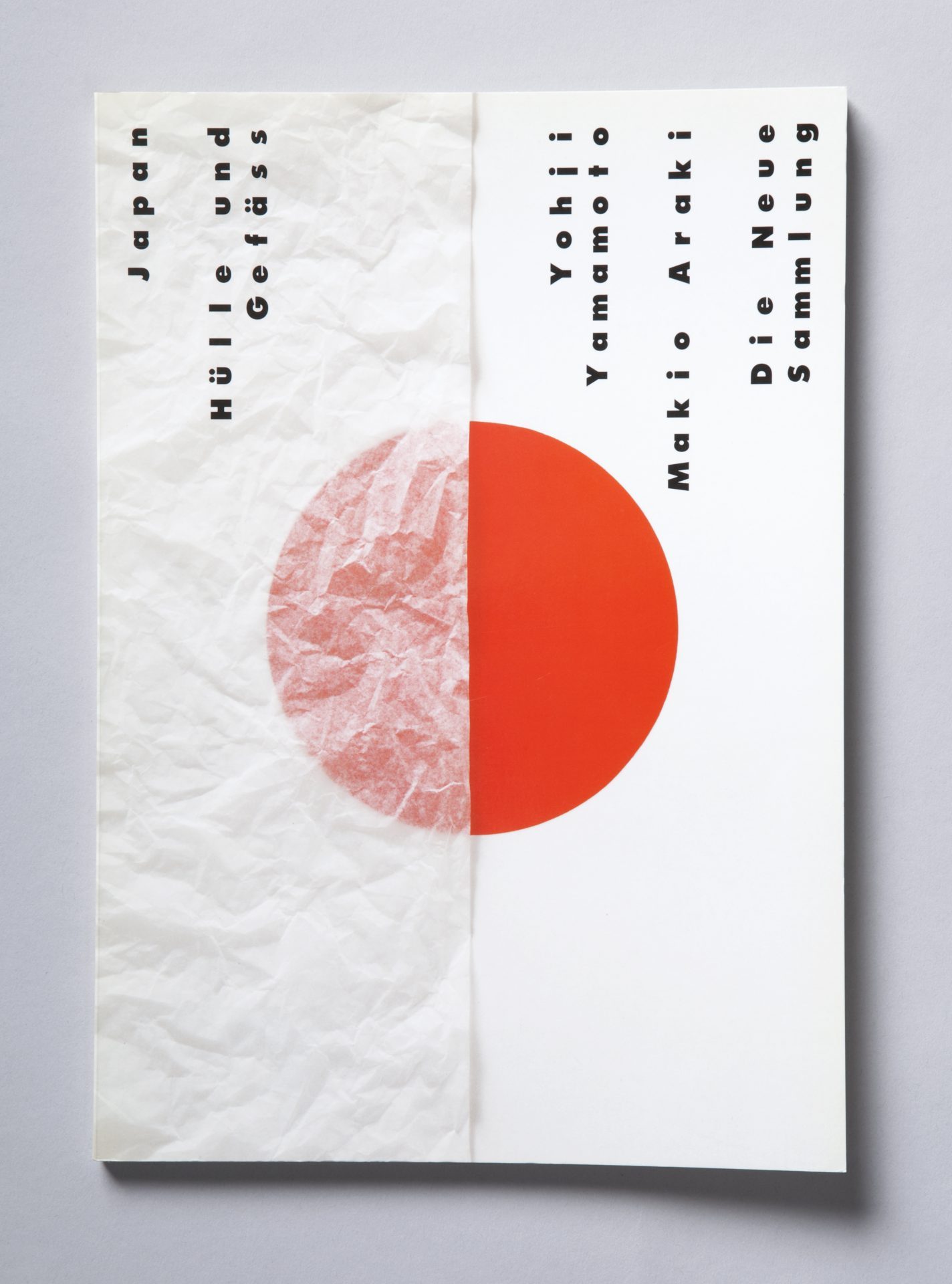 Zweigeteiltes weißes Cover mit schwarzer Beschriftung, darunter mittig großer roter Punkt, der senkrecht von einem bis in die Mitte reichenden, weiß-transparentem Papier überdeckt ist. Beschriftung, senkrecht: von unten nach oben: Japan Hülle und Gefäß Yohji Yamamoto Makio Araki Die Neue Sammlung.