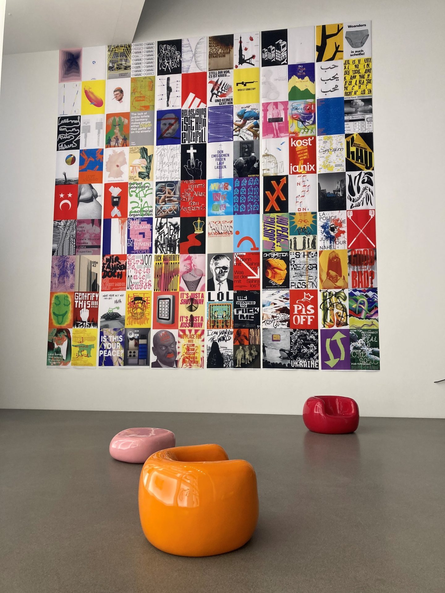 Photo of the wall with posters. A total of 108 colourful posters are arranged in a rectangle. In front of them are three armchairs in orange, pink and red.