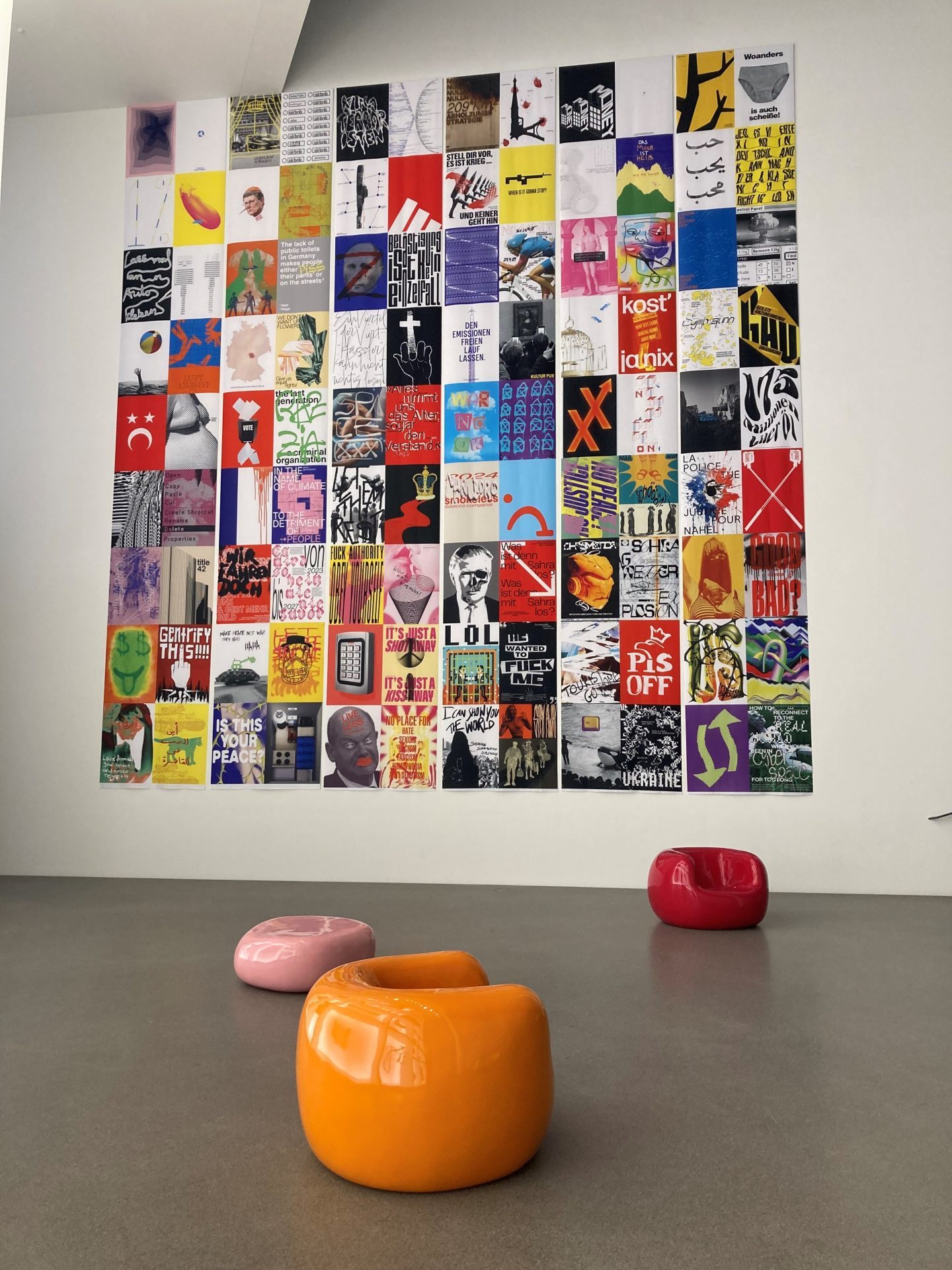 Foto von der Wand mit Postern. In einem Rechteck sind insgesamt 108 bunte Poster angeordnet. Davor befinden sich drei Sessel in orange, rosa und rot.